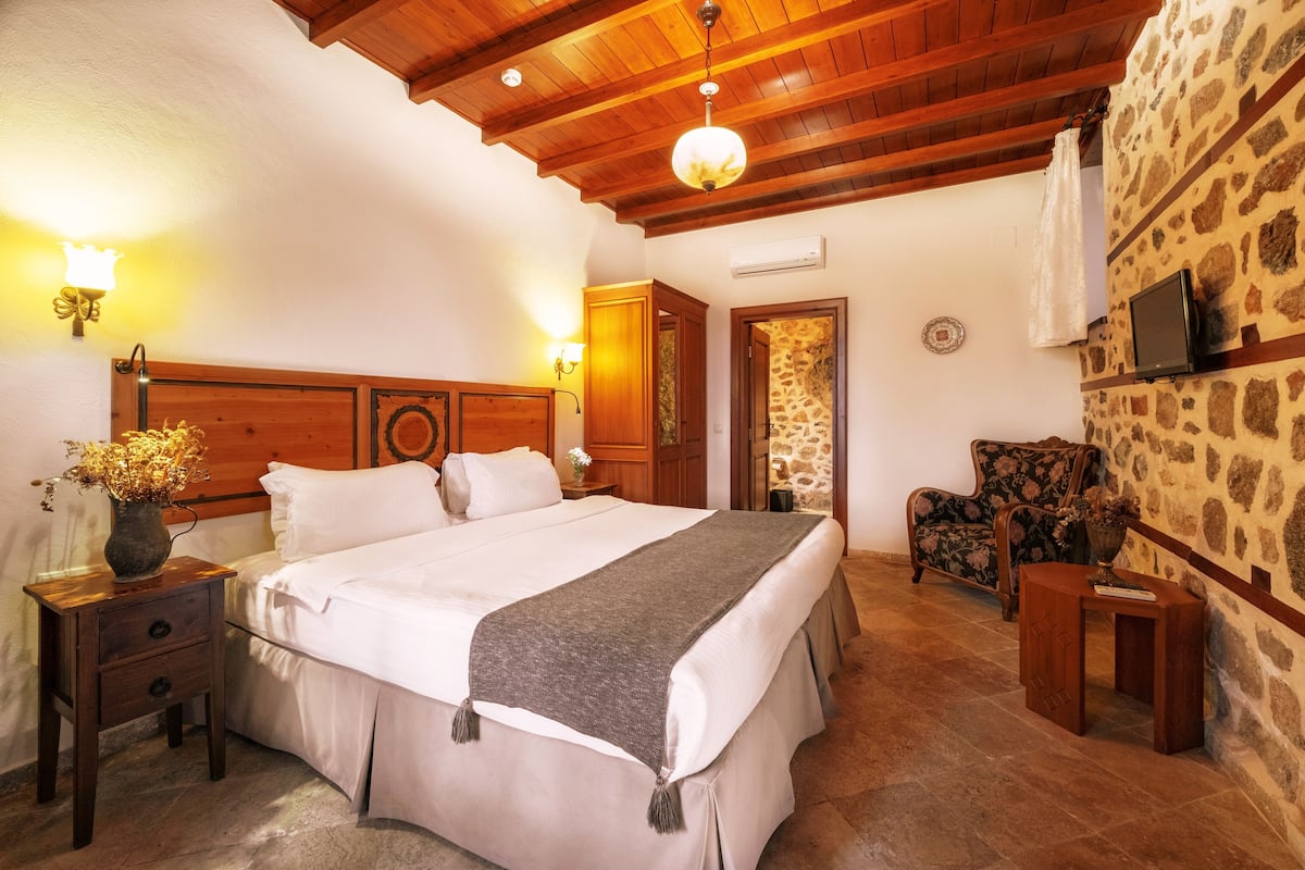 A bedroom with a wooden headboard, a bed with white bedding, a nightstand with a vase of flowers, a chair, and a stone wall.