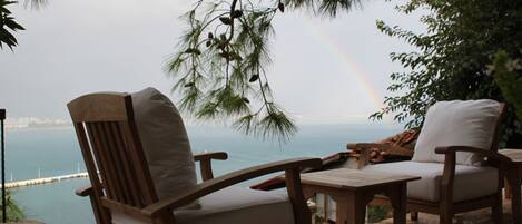 Un espacio de estar al aire libre con sillas y una mesa de madera, con vistas a un cuerpo de agua y un arcoíris.