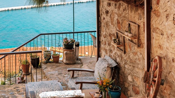 A stone-walled balcony with a view of the sea, wooden furniture, potted plants, and a rustic wheel. - Hotel Villa Turka (Alanya)