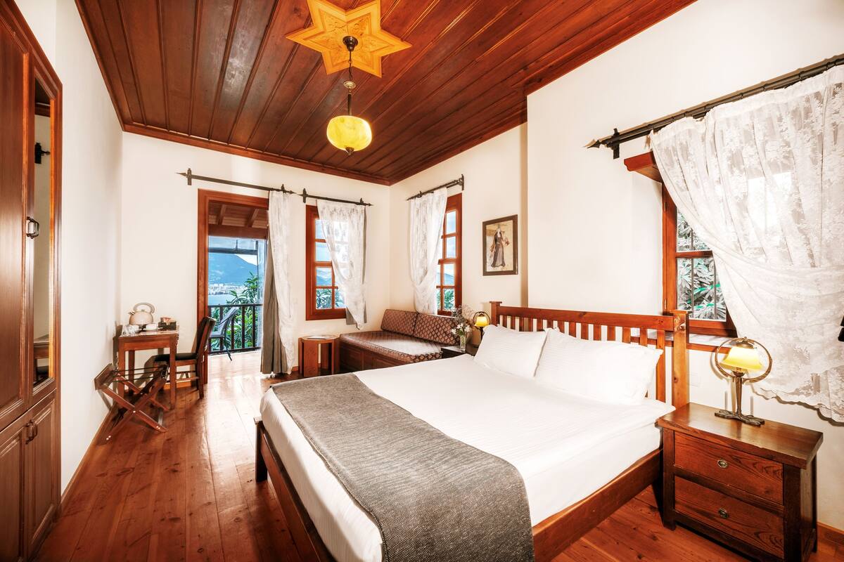 A bedroom with a wooden ceiling, a large bed, a bedside table with a lamp, a window with curtains, and a view of the outdoors.
