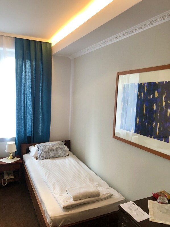 Ein Hotelzimmer mit einem Bett, einem Nachttisch mit Lampe, einem Telefon, einem Bilderrahmen mit abstrakter Kunst und einem Fenster mit blauen Vorhängen.