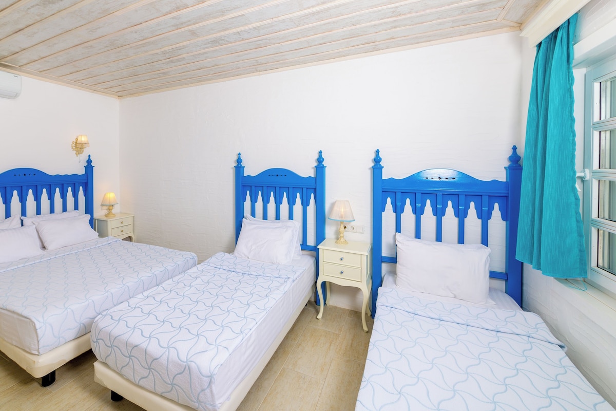 A room with three beds, each with a blue headboard and white bedding. There are bedside tables with lamps, a ceiling fan, and a window with curtains.