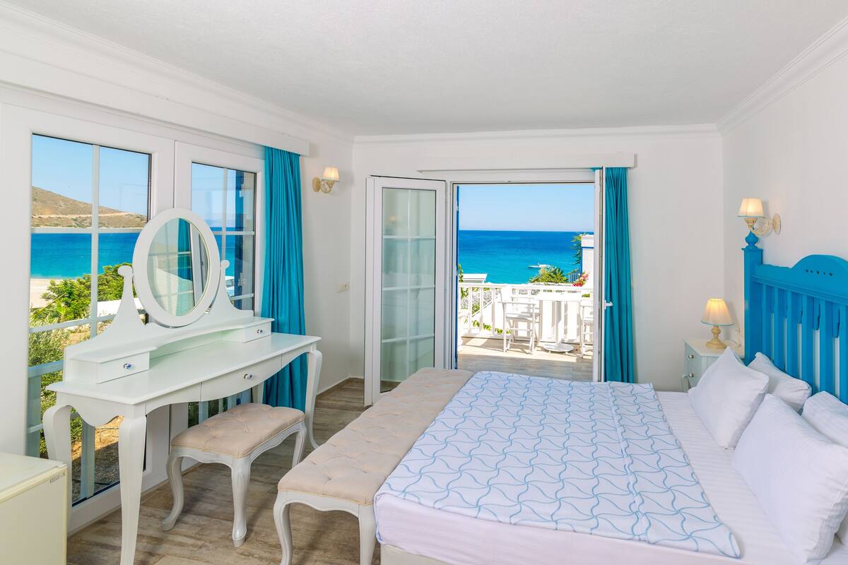 A bedroom with a bed, a vanity with a mirror, a desk, and a view of the sea.
