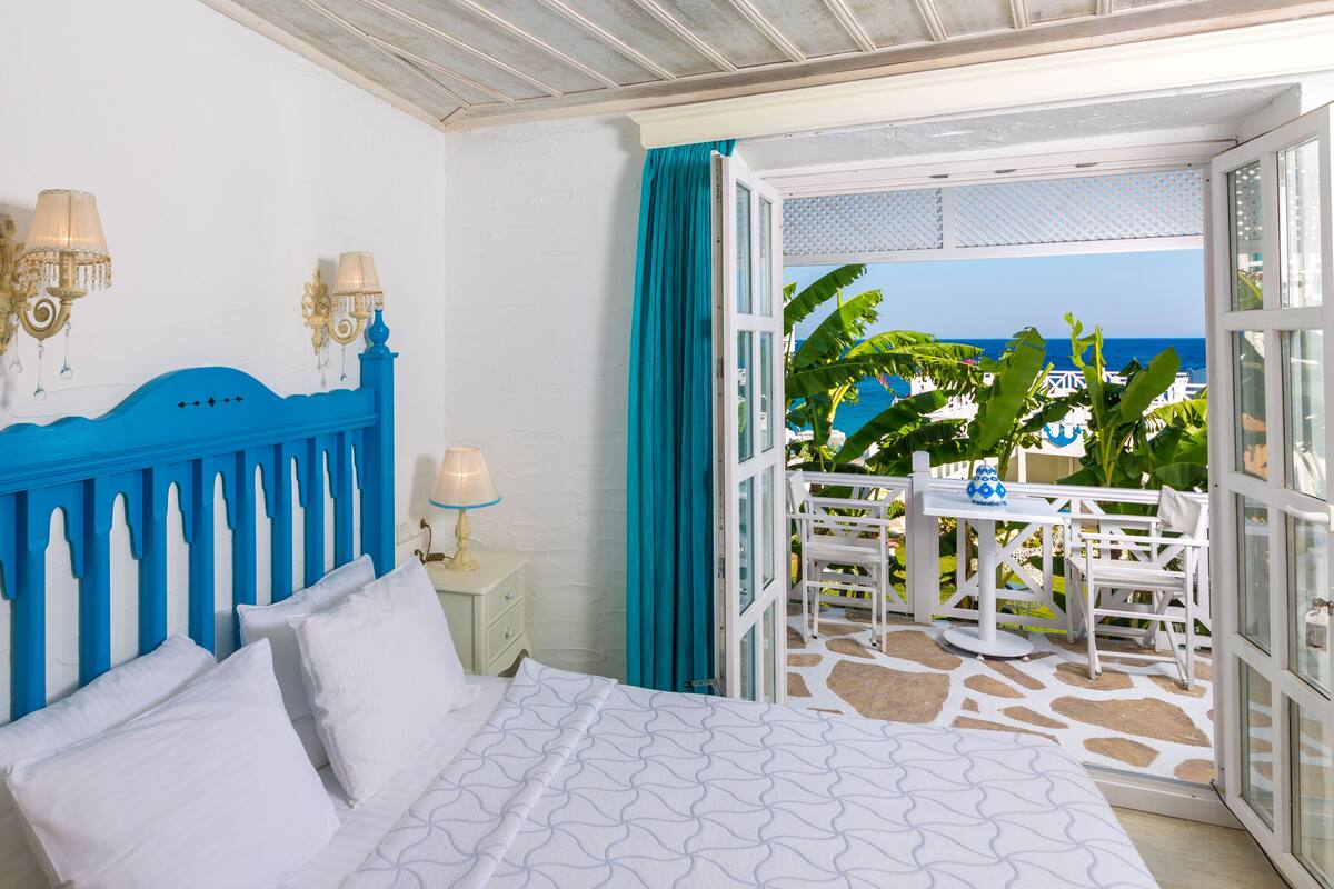A bedroom with a blue headboard, white bedding, and a view of the ocean through an open door.