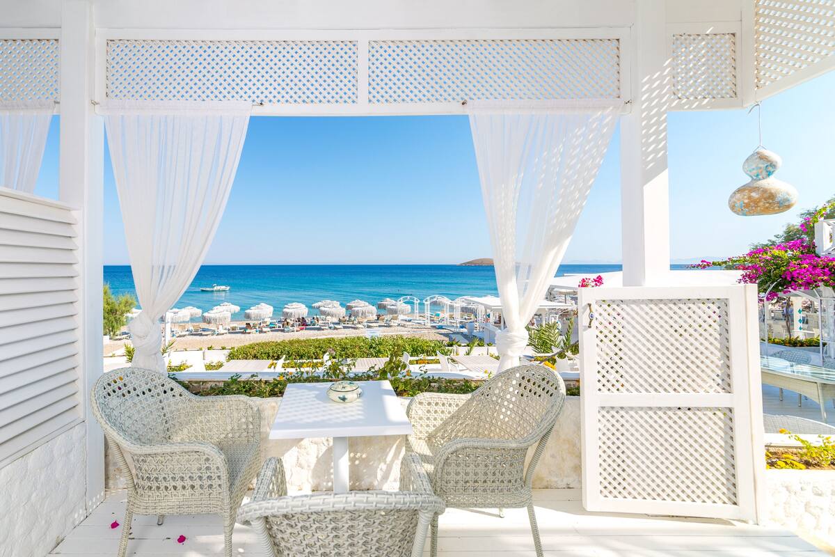 A covered outdoor seating area with wicker chairs and a table, overlooking a beach with umbrellas and a clear blue sky.