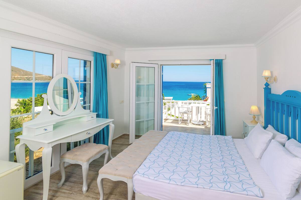 A bedroom with a bed, a vanity with a mirror, a desk, and a view of the sea.