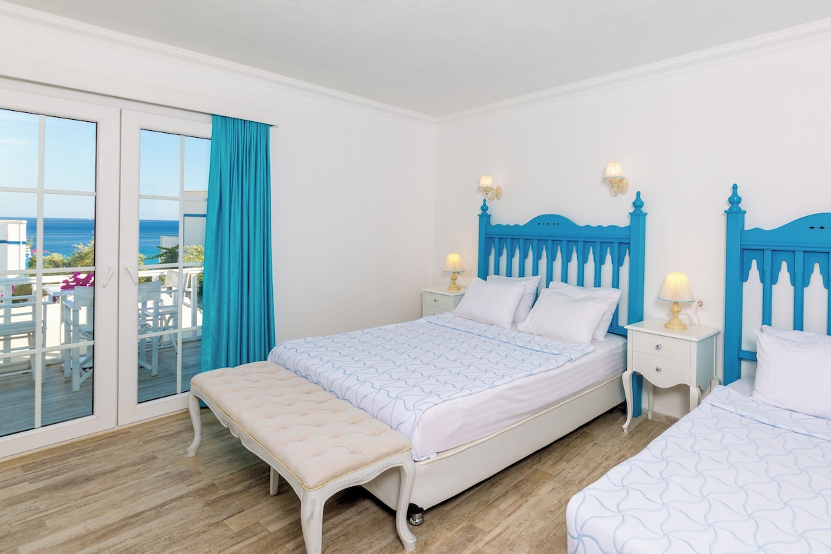 A bedroom with a large bed, a smaller bed, a bench, two bedside tables with lamps, and a view of the sea through a sliding door.