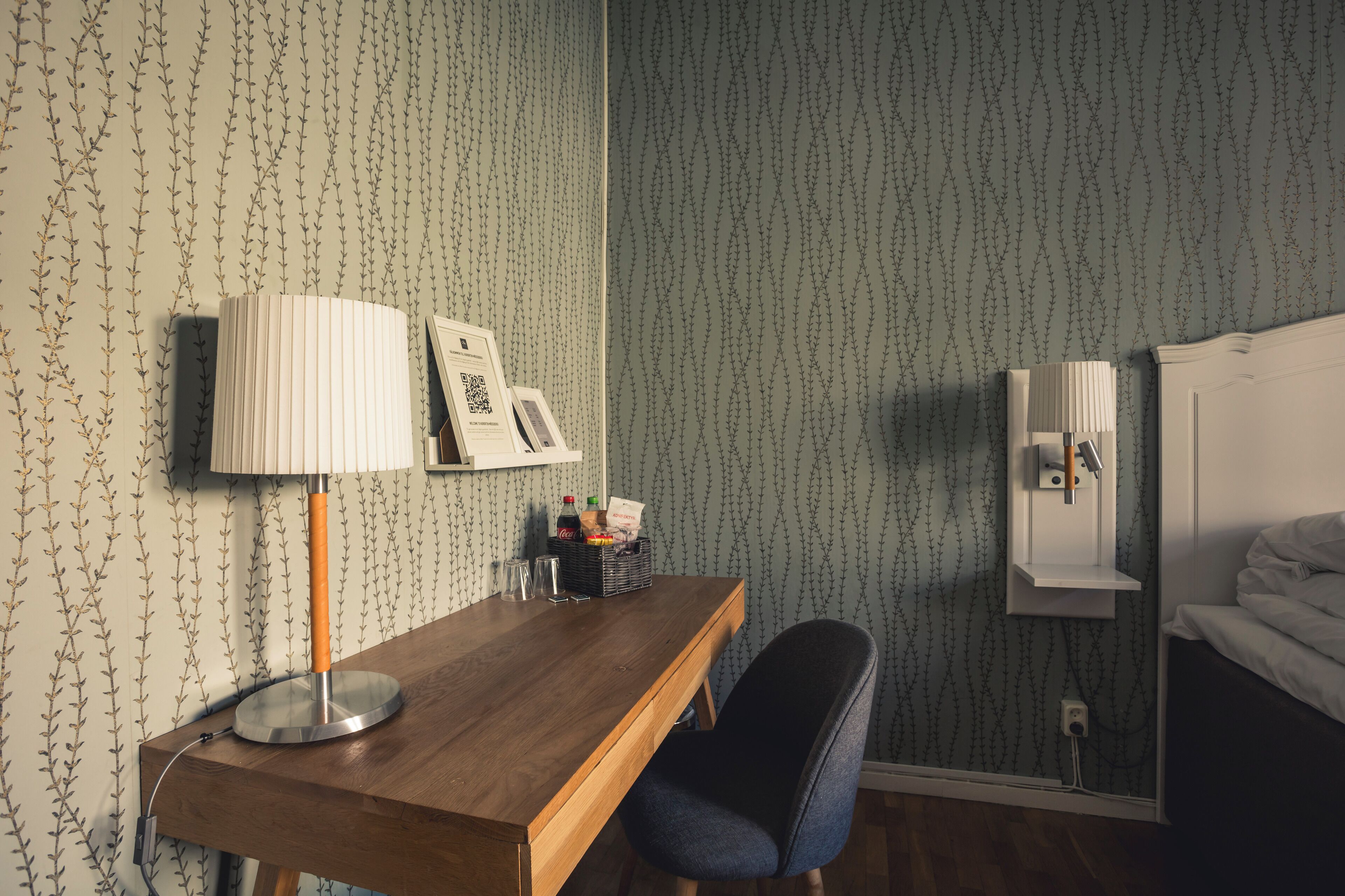 A hotel room with a wooden desk, a lamp, a chair, a bed, and a wall-mounted shelf with a mirror and a picture frame.