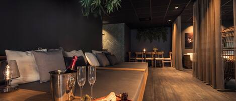 A modern hotel room with a wooden bench, a minibar, and a seating area with a sofa and chairs.