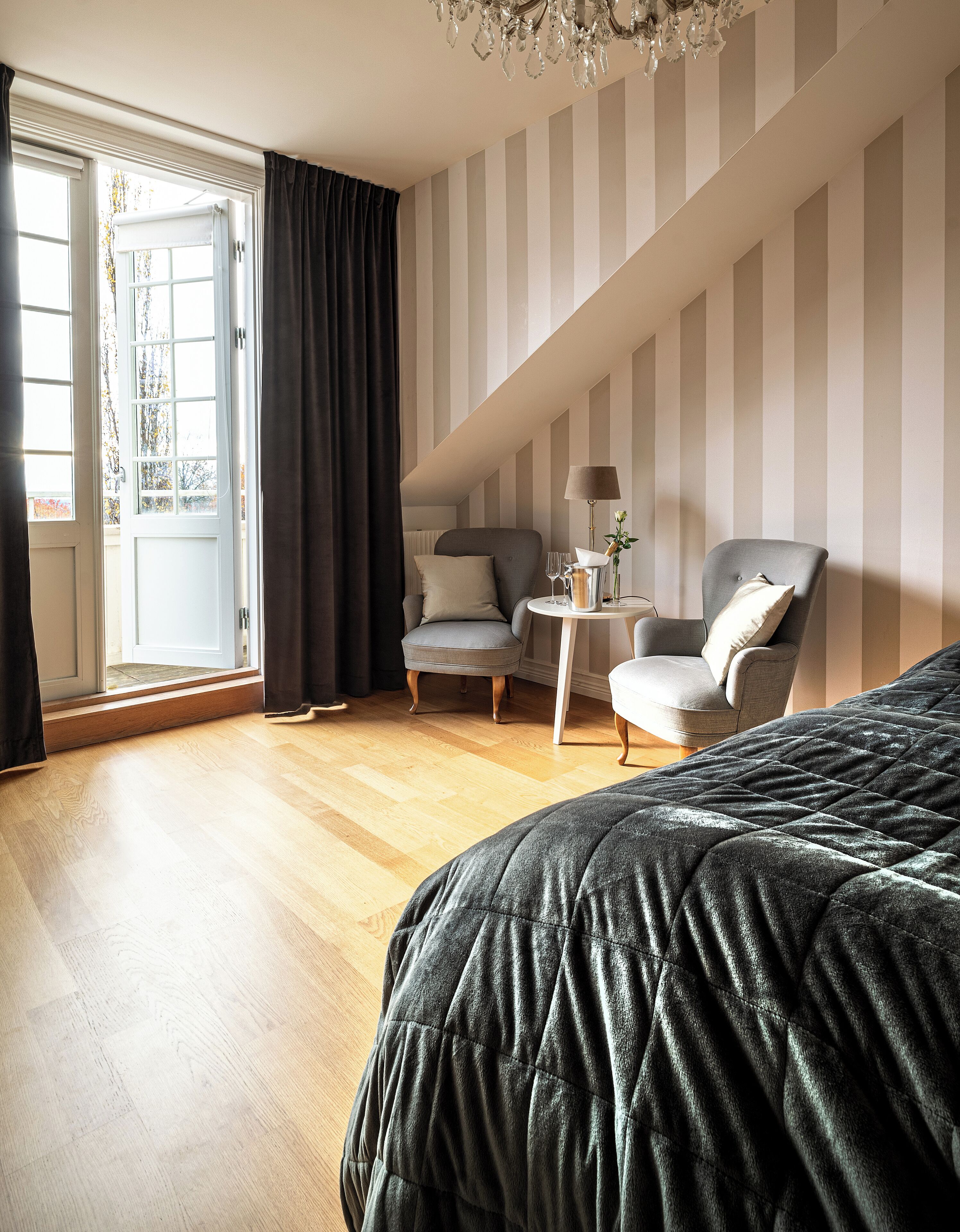 A modern hotel room with a large bed, two armchairs, a small table with a lamp, and a view of the outdoors through a large window.