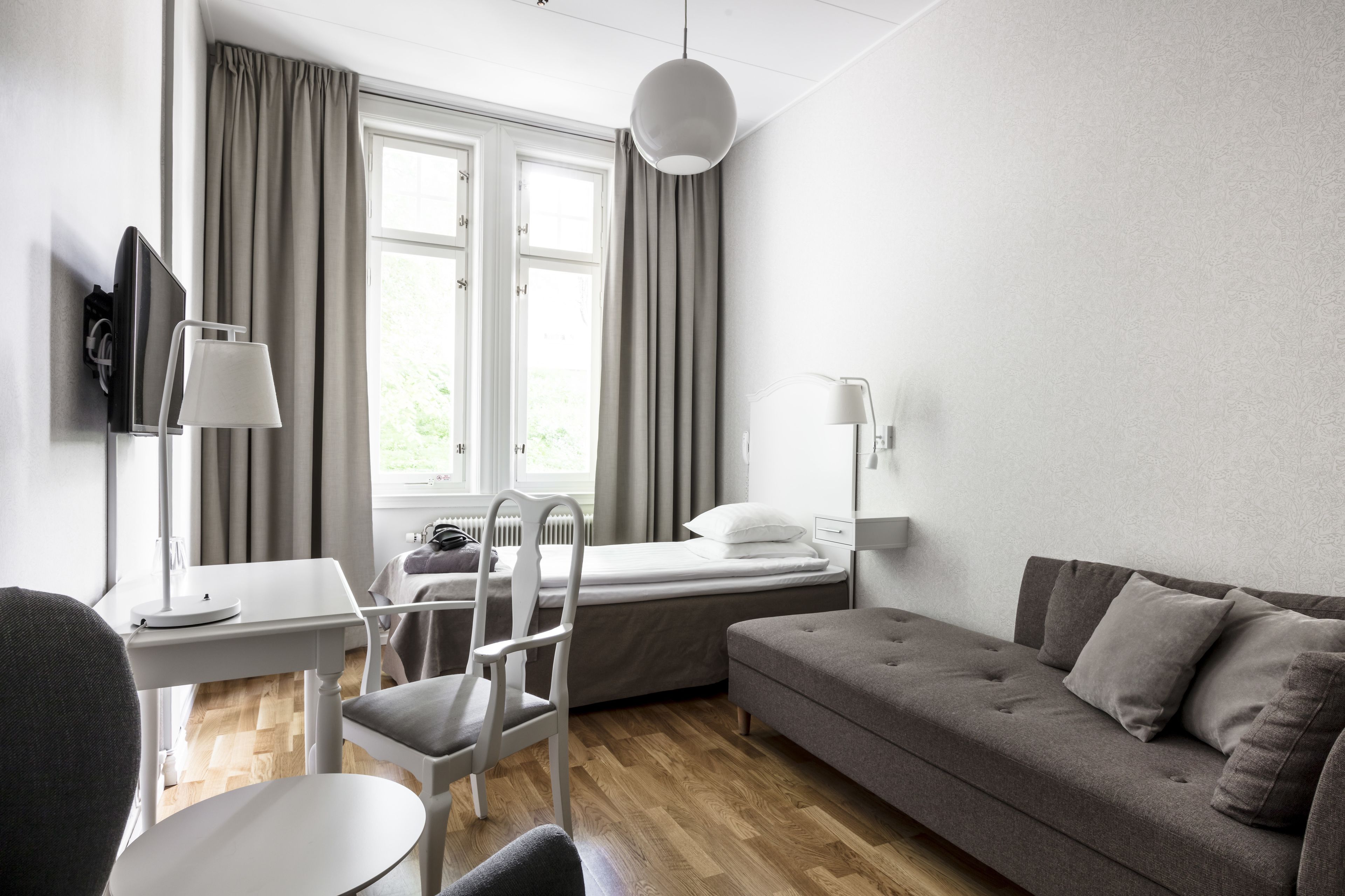 Una habitación moderna y luminosa, con piso de madera, un sofá gris, una silla blanca, una mesita y una cama con cabecera blanca.