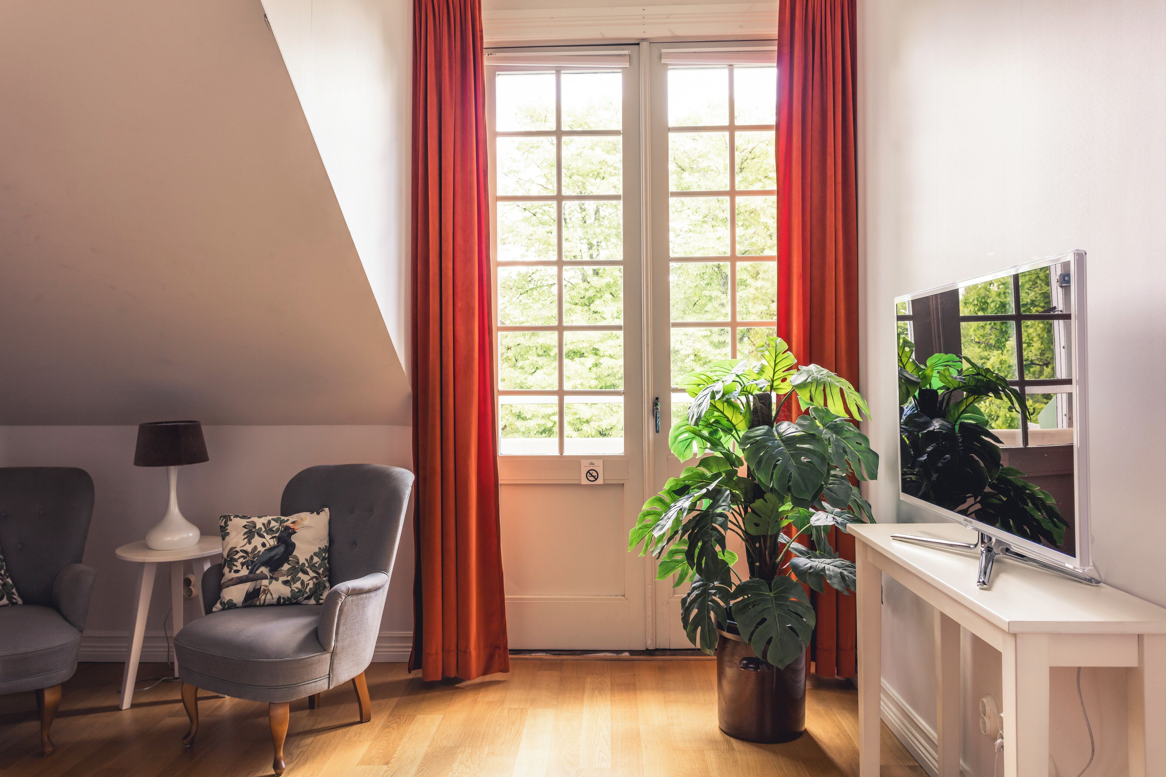 Une pièce comprenant deux fauteuils gris, une table d’appoint blanche avec une lampe, une grande plante en pot, une cheminée blanche et un téléviseur fixé au mur.