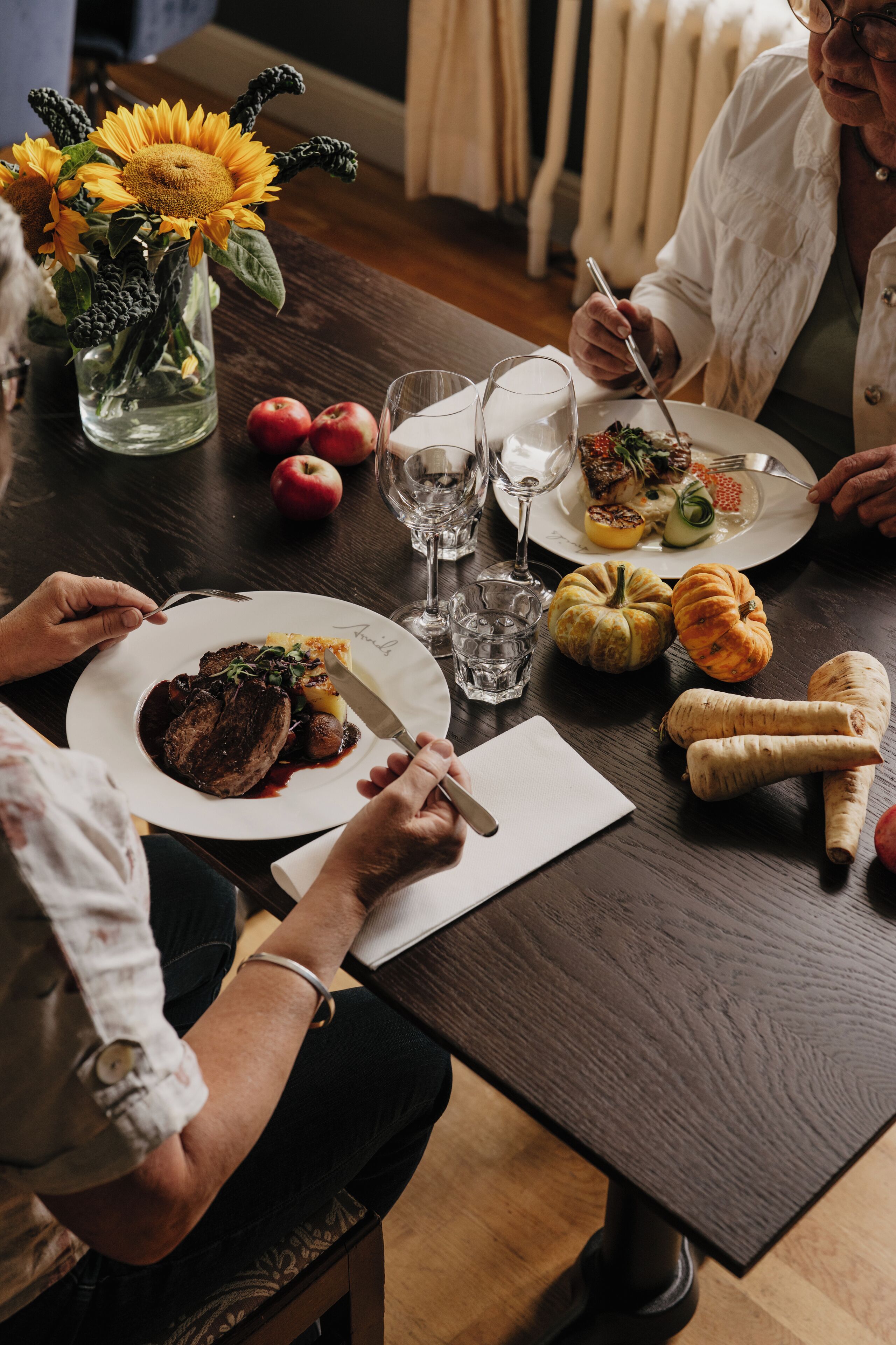 A dining table with a meal, a vase with sunflowers, and a person writing.
