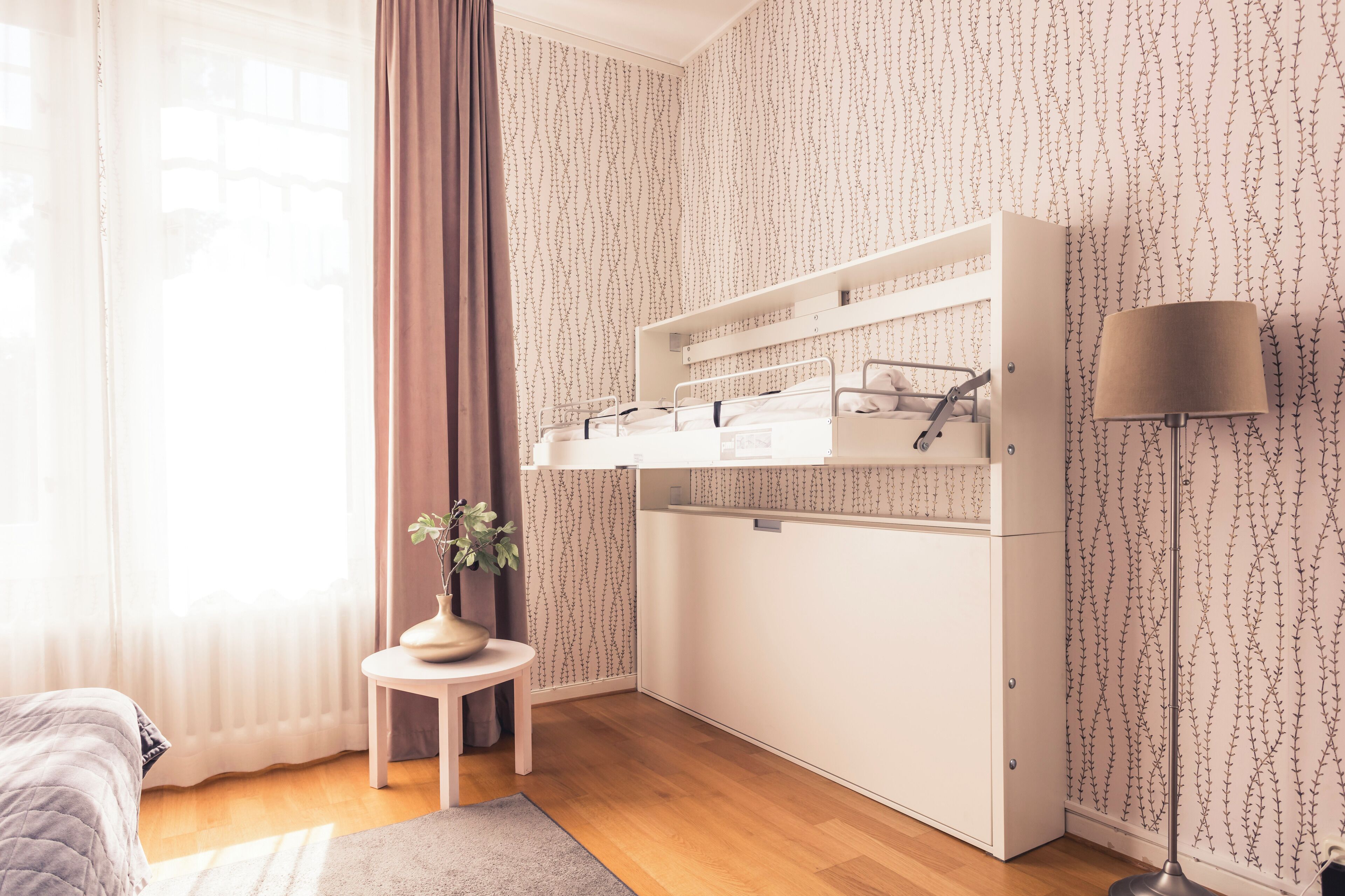 Un dormitorio con una cama individual sobre otra, una mesita de noche con una planta, una lámpara y una ventana con cortinas.