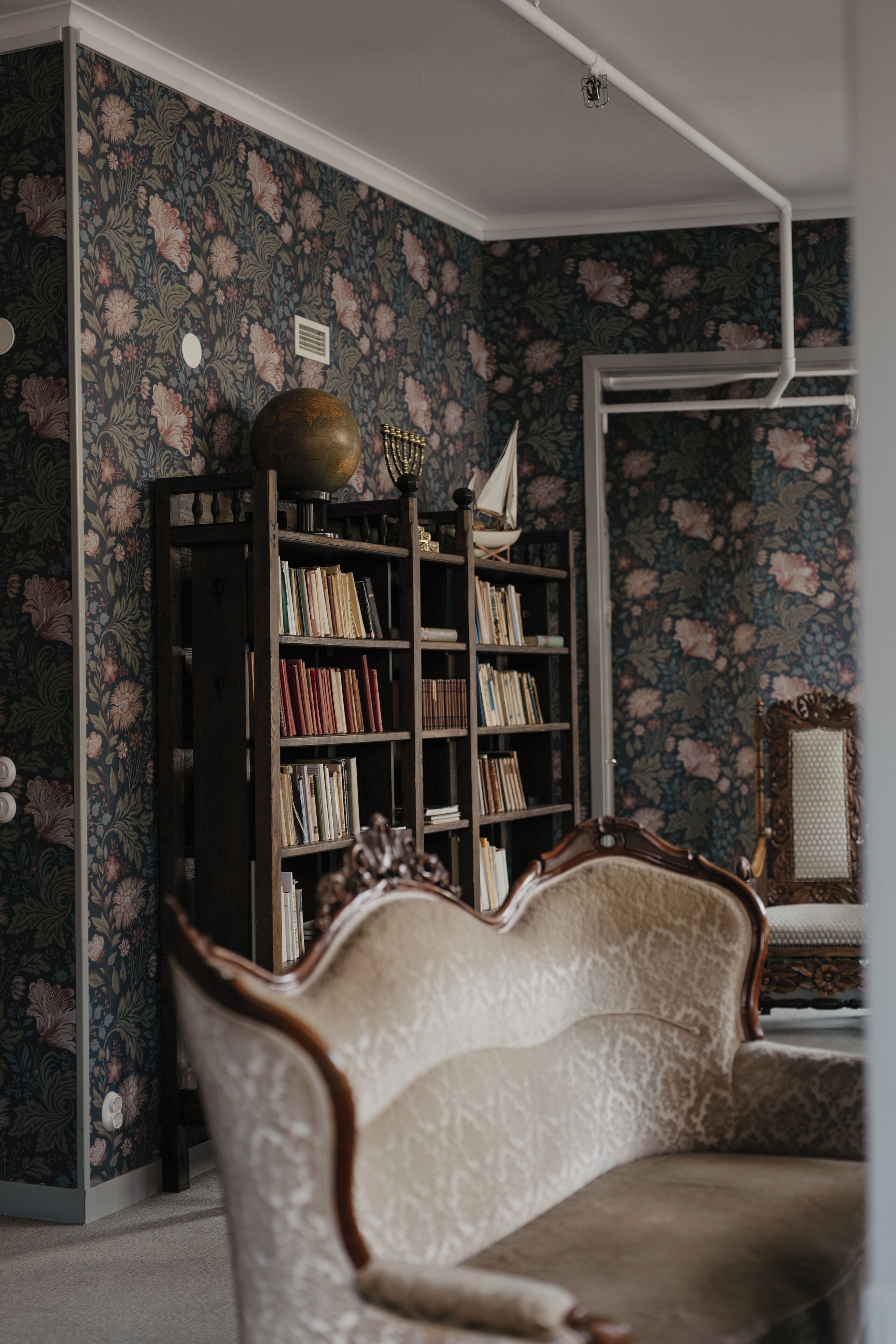 A room with a floral wallpaper, a bookshelf, and a vintage armchair.