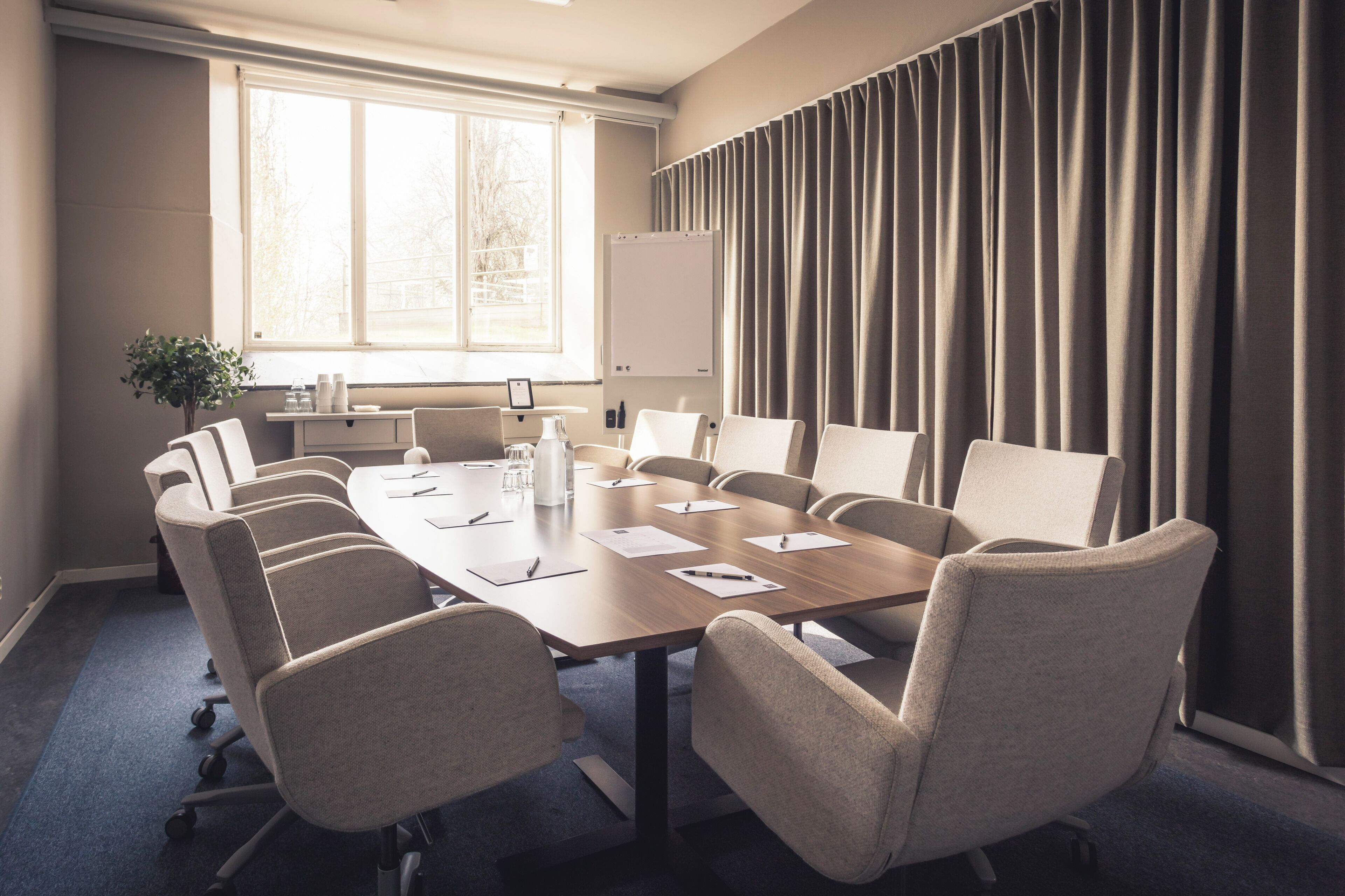 A conference room with a long table, multiple chairs, a whiteboard, and a window with curtains.