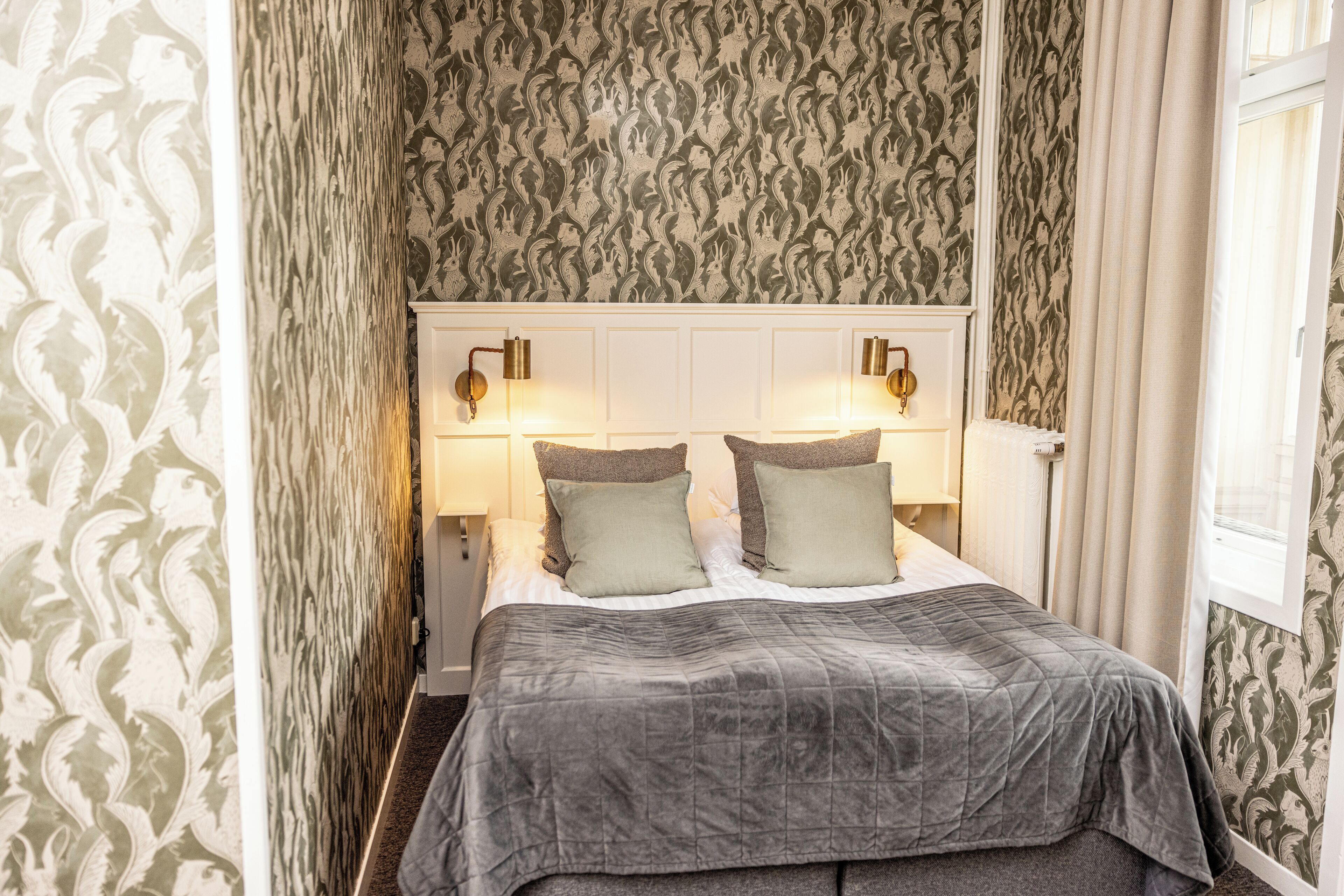 A neatly made bed with a grey quilt, two green pillows, and a white headboard with wall-mounted lamps.