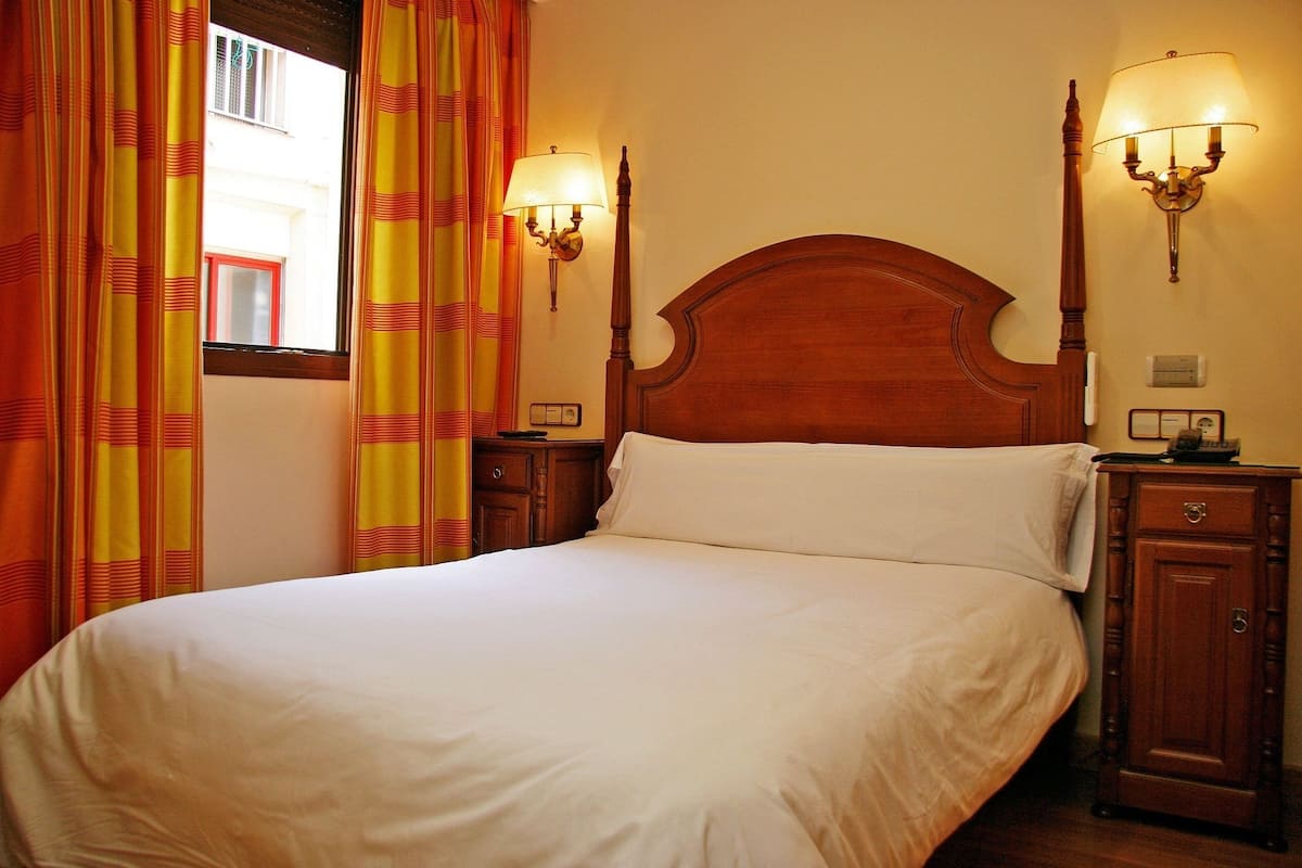 A neatly made bed in a room with a wooden headboard, two wall-mounted lamps, and curtains.