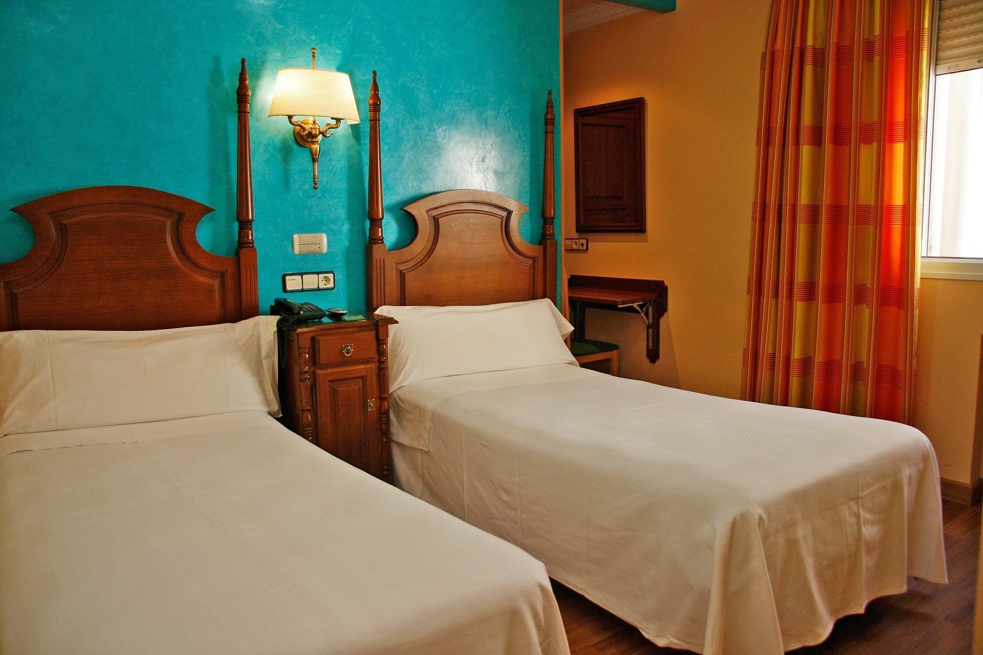 A hotel room with two single beds, a nightstand with a lamp, a small table, and a window with curtains.