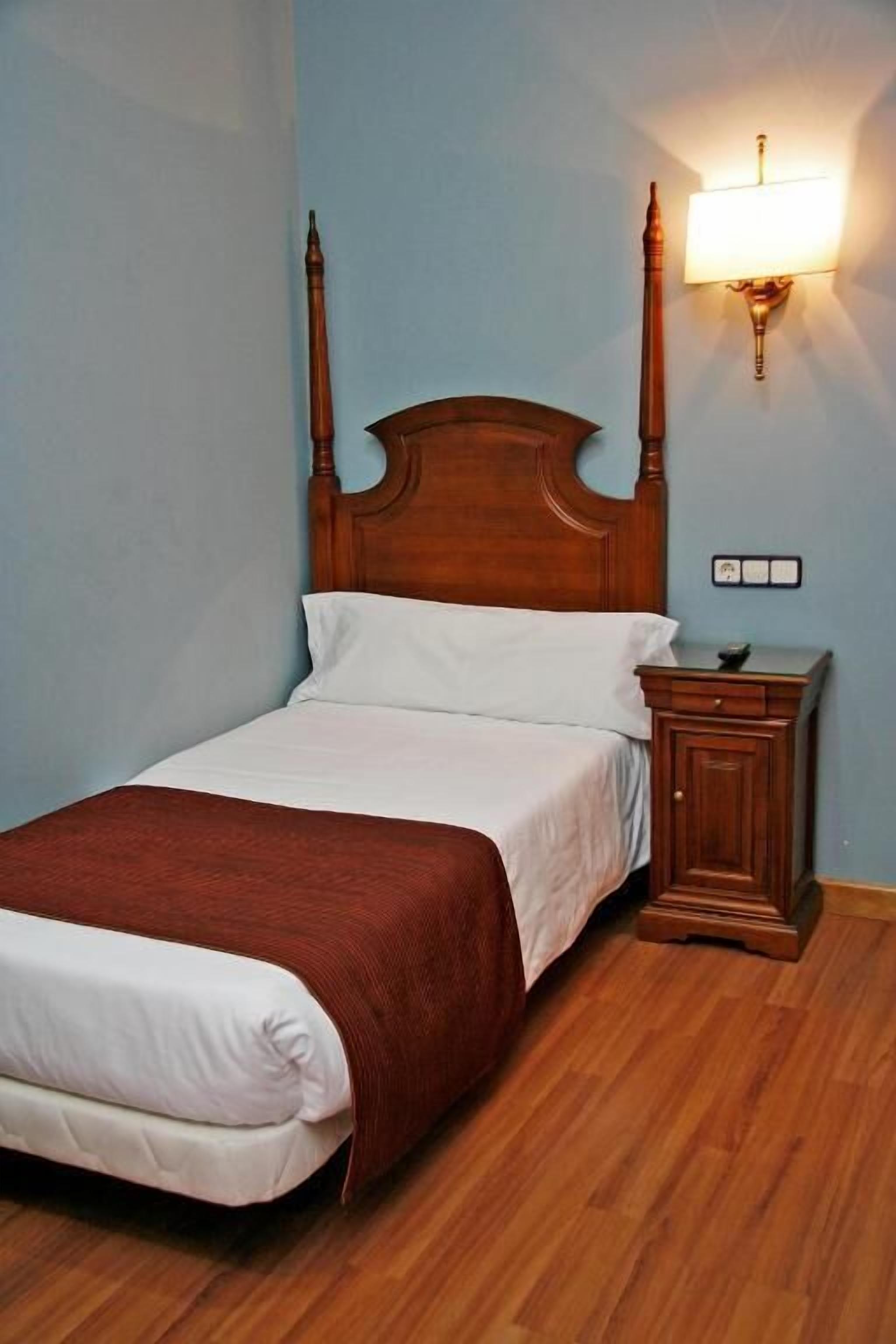 Una cama bien tendida con cabecera de madera, una mesita de noche con lámpara y un interruptor de pared.