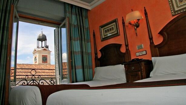 Hotelkamer met een groot bed, nachtkastje, lamp en uitzicht op een toren door een open raam.
