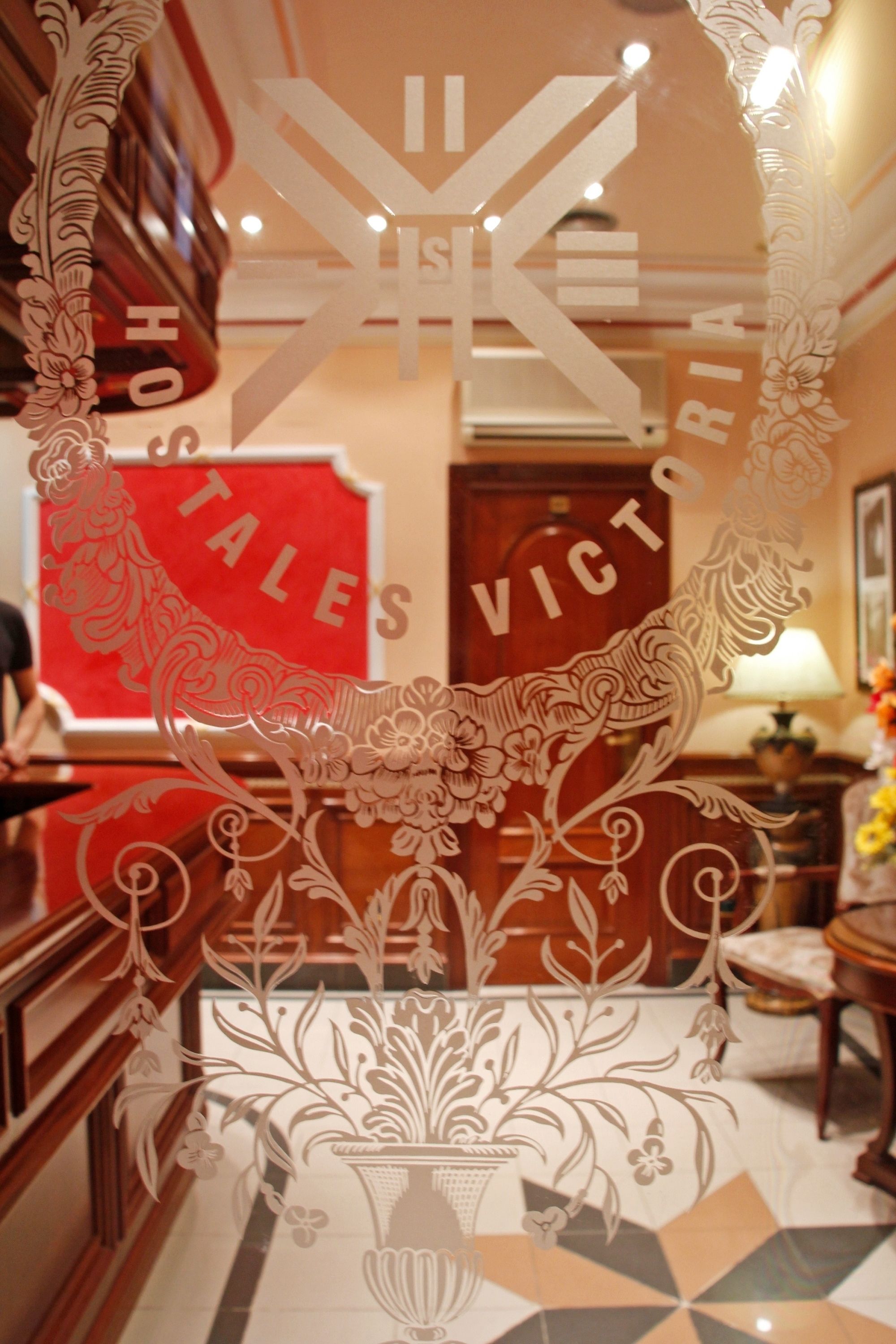 A hotel lobby with ornate white paper cutouts, a red sign reading "HO HO TALES VICTORIA," and a patterned floor.