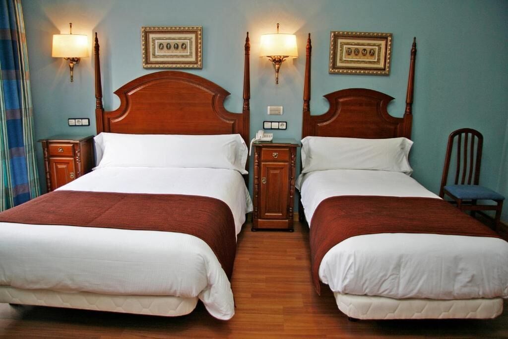 A hotel room with two double beds, wooden headboards, bedside tables with lamps, and framed pictures on the wall.