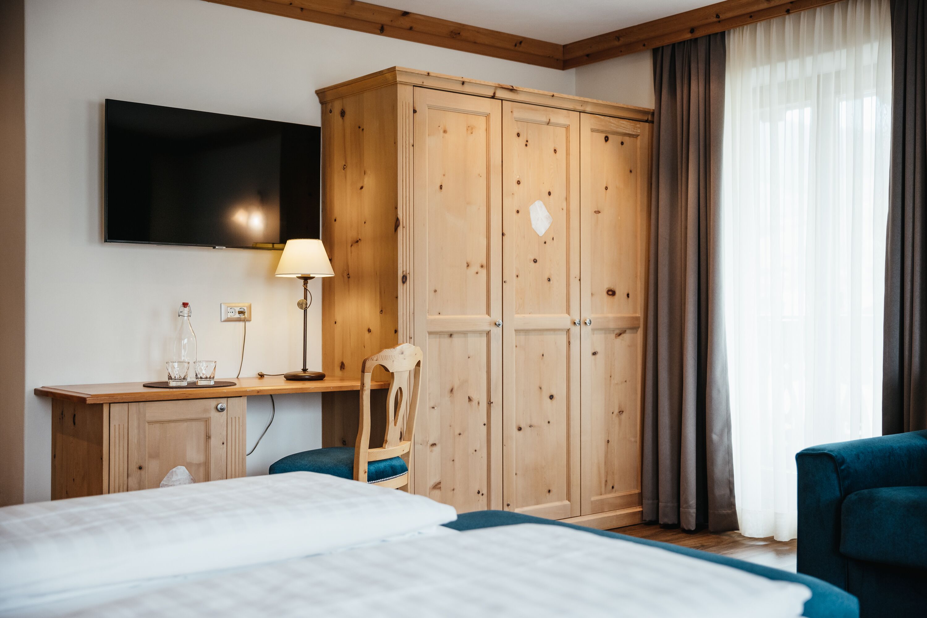 Habitación de hotel con un armario de madera, un televisor montado en la pared, un escritorio con lámpara, dos camas y una silla.
