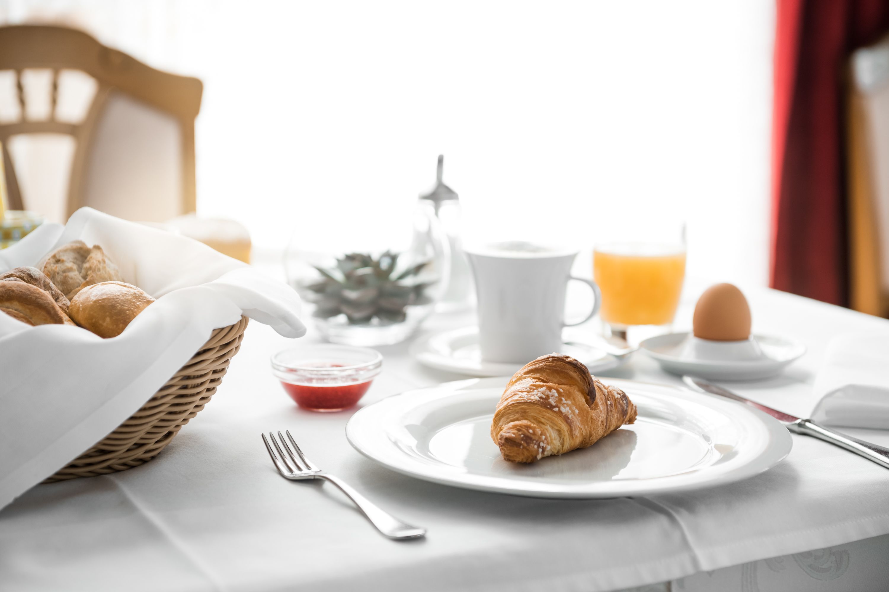 Ein Frühstückstisch mit Croissants, einer Tasse Kaffee, einem Glas Orangensaft und einer kleinen Schale Marmelade auf einer weißen Tischdecke.