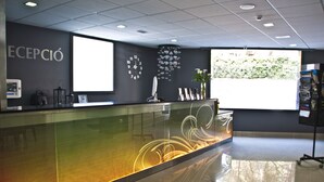 A reception area with a glass counter, a clock, and a large window with a view of greenery. - Summer Hotel (Calella)