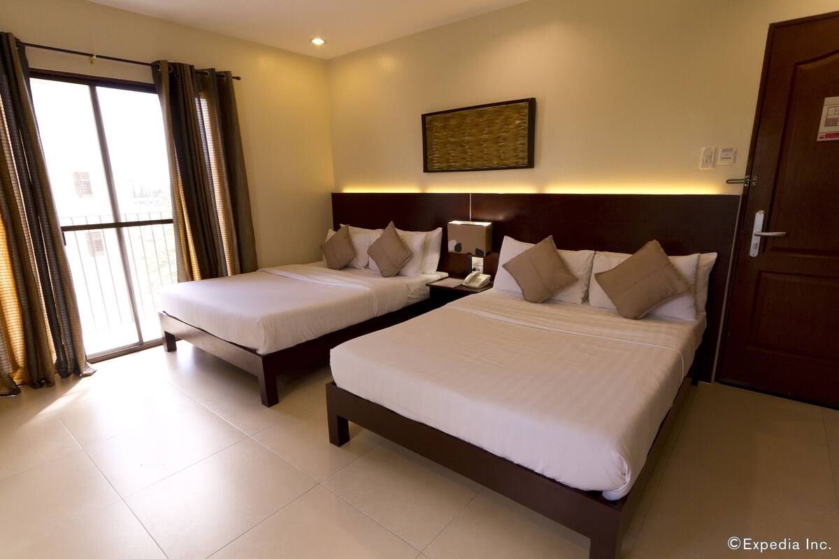 A hotel room with two beds, a large window with curtains, a wall-mounted artwork, and a door.