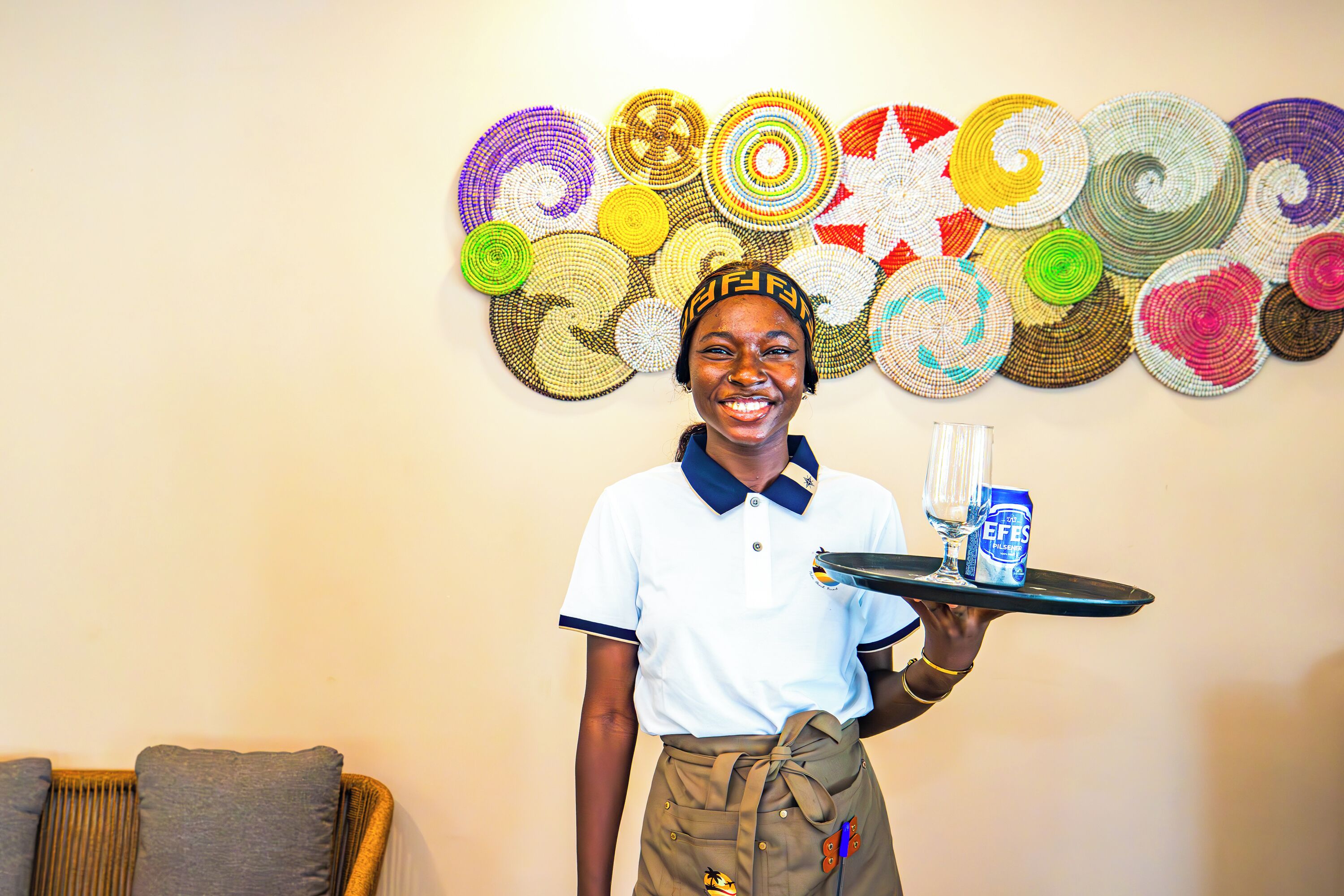 A person holding a tray with a glass and a can, standing in front of a wall with colorful woven art.