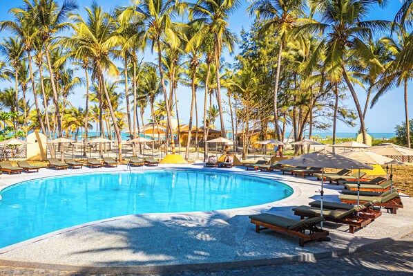 A swimming pool surrounded by lounge chairs and umbrellas, with palm trees and a view of the ocean. - Palm Beach Resort (Serrekunda)