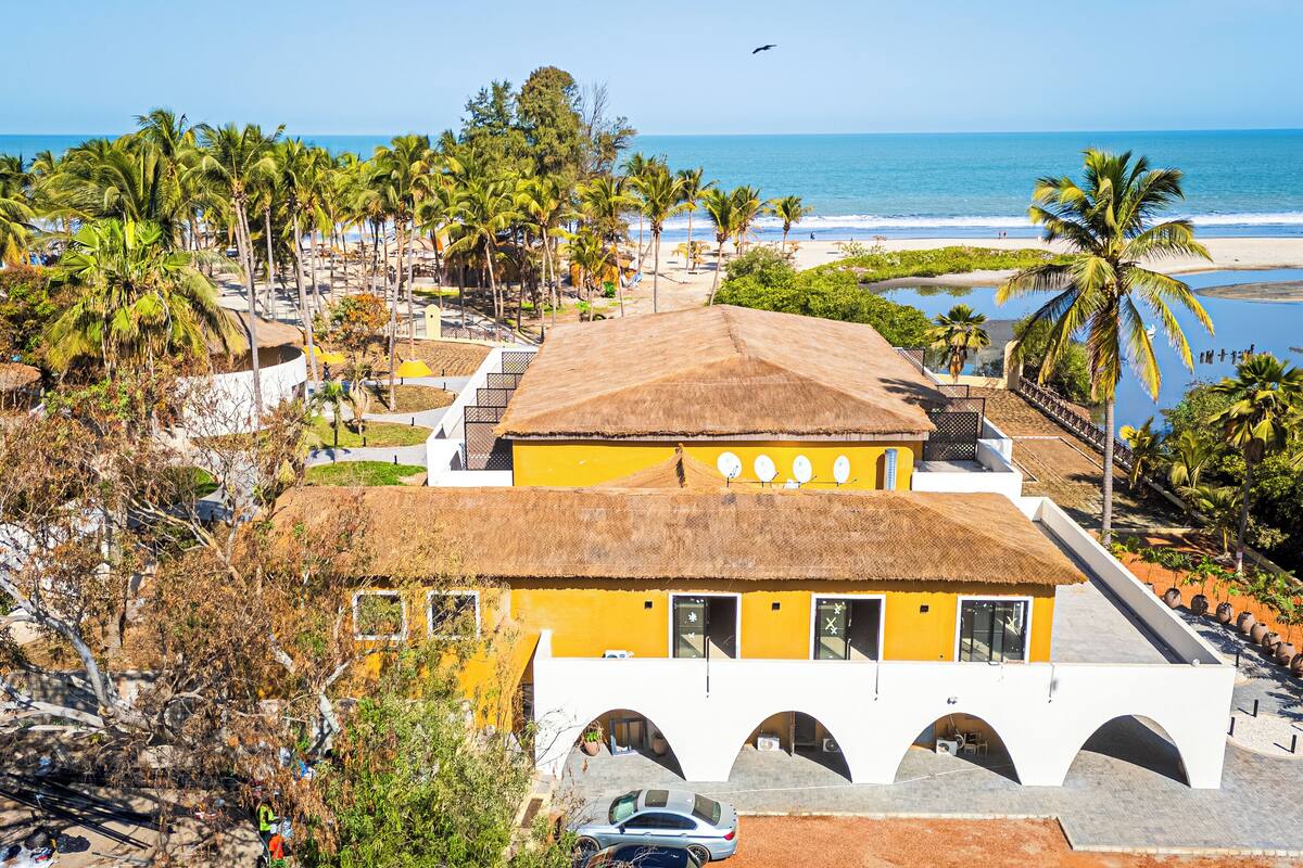 A building with a thatched roof, white walls, and arched windows, surrounded by palm trees and a beach.