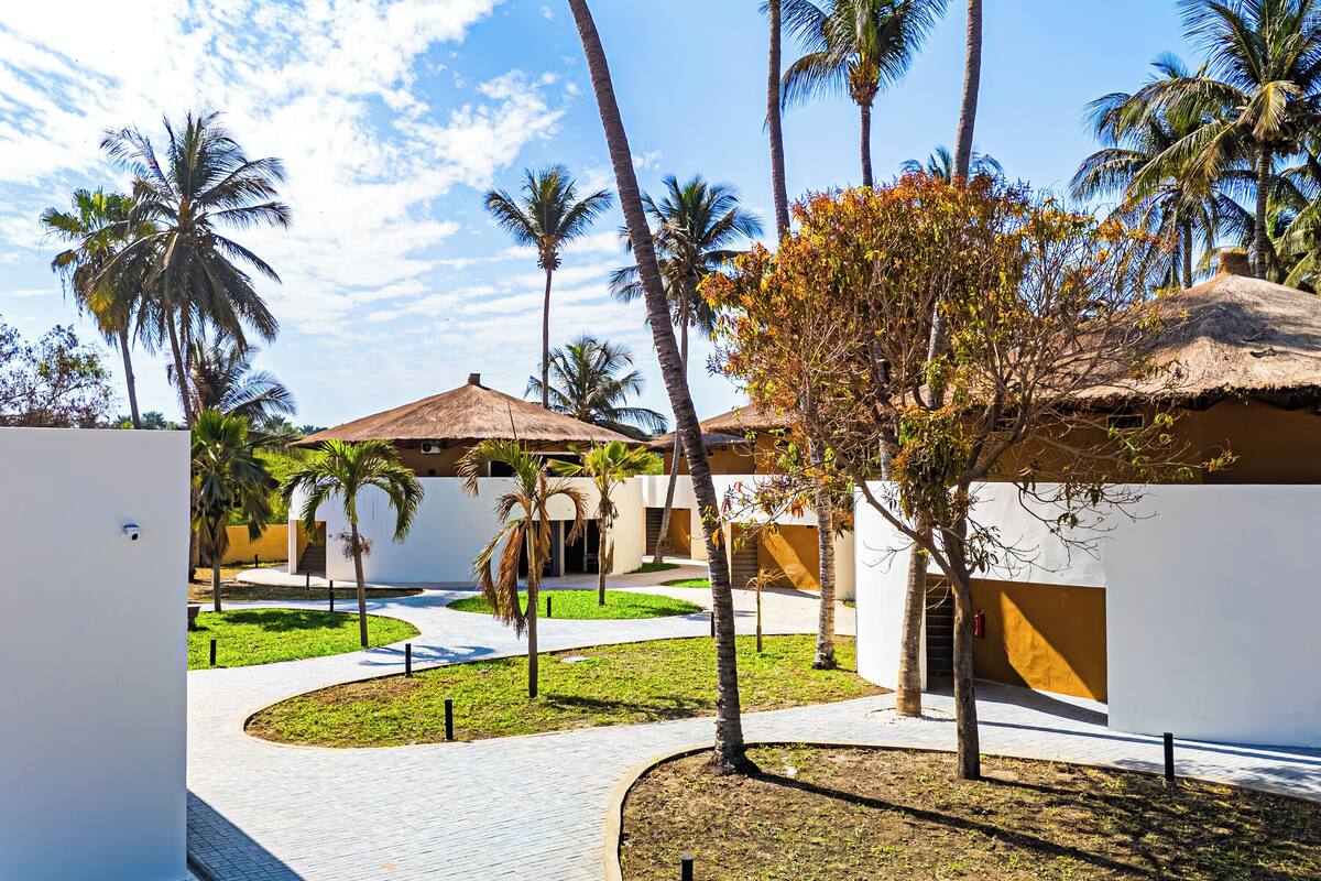 A courtyard with palm trees, a thatched-roof building, and a white wall.