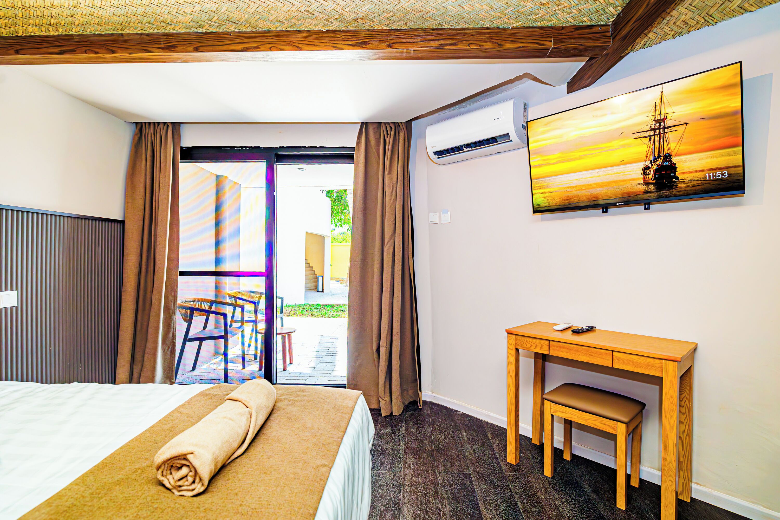 A hotel room with a bed, a desk, a chair, an air conditioning unit, and a TV displaying a sunset scene.