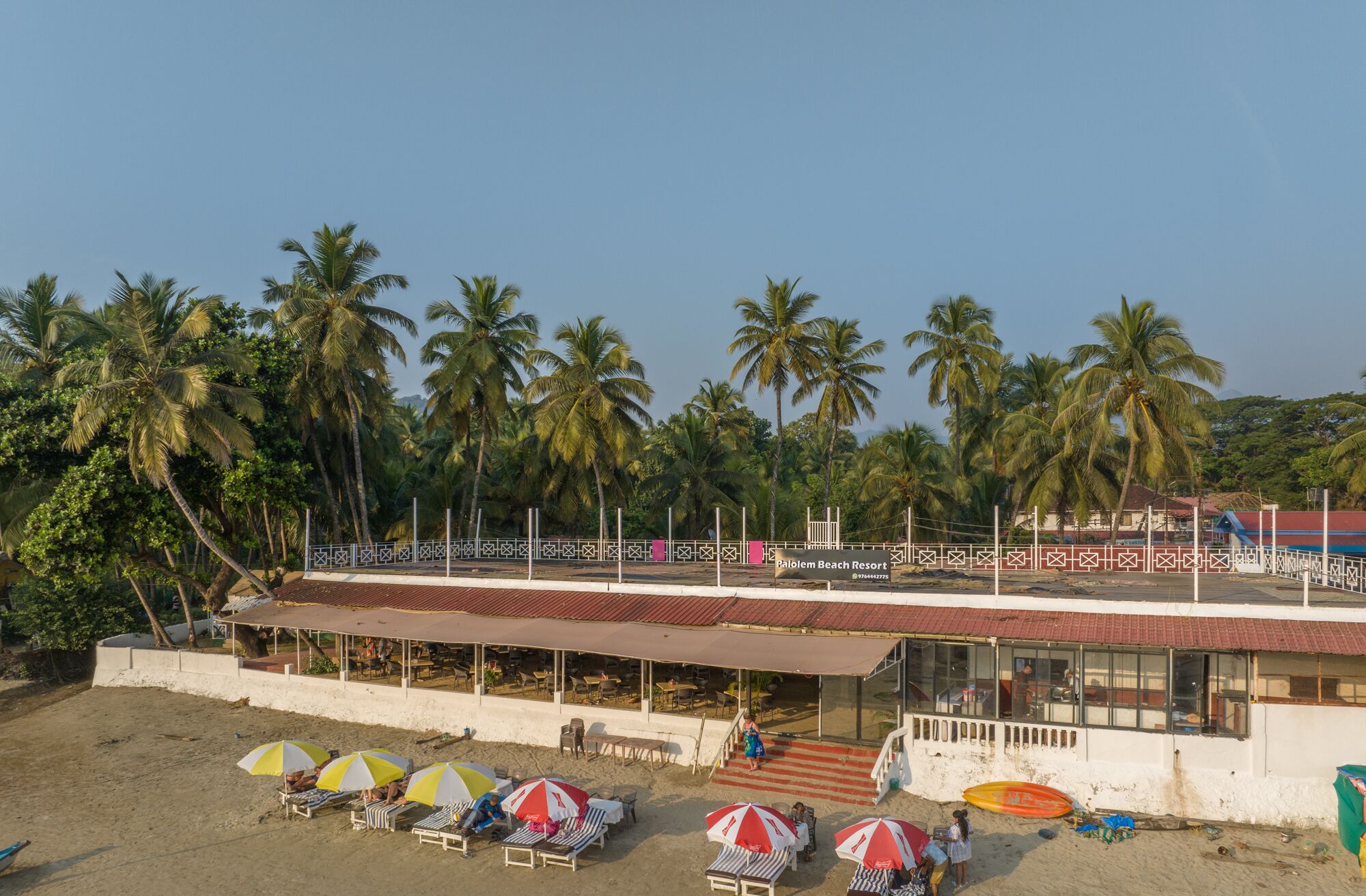 Un resort en la playa con un área de comedor cubierta, sillas de playa, sombrillas y vistas a palmeras.