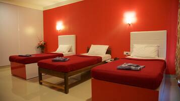 Una habitación con tres camas, cada una con una colcha roja y cabecero blanco. Hay una mesita entre las camas y una lámpara de pared sobre cada cama.