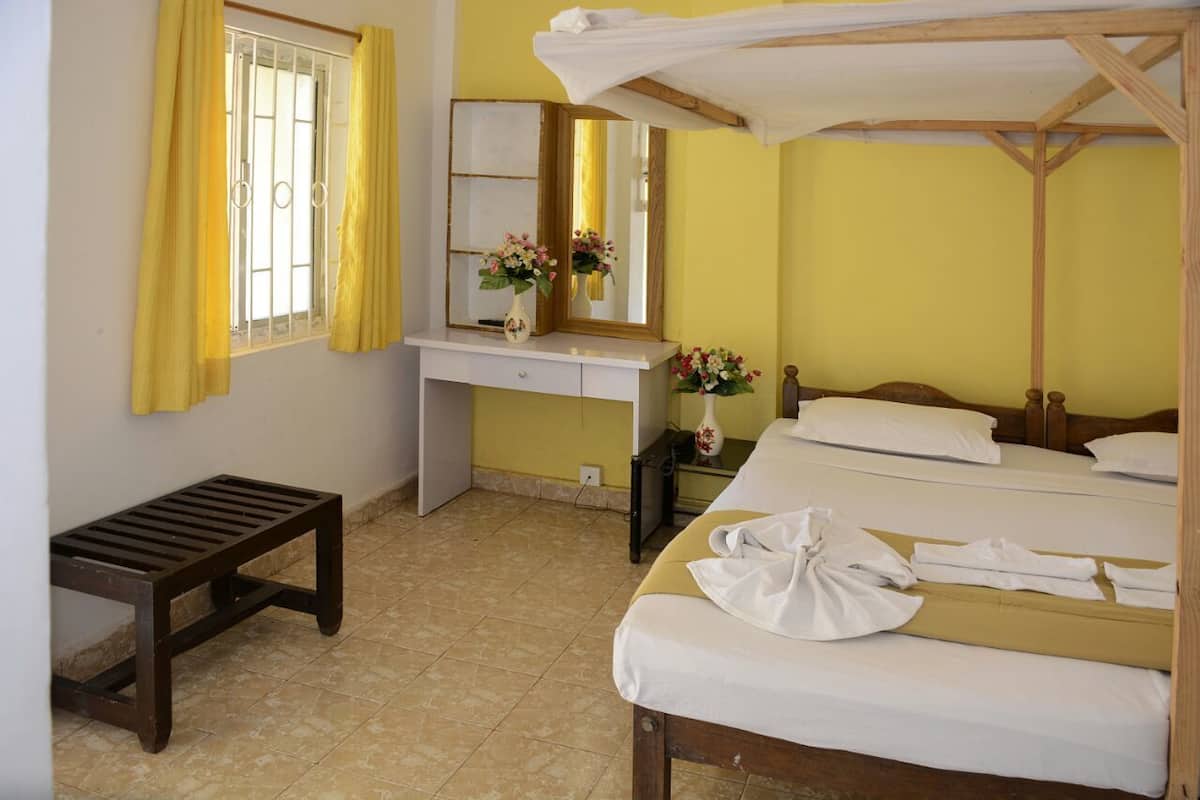 Una habitación de hotel con cama, mesita de noche, escritorio con espejo y ventana con cortinas amarillas.