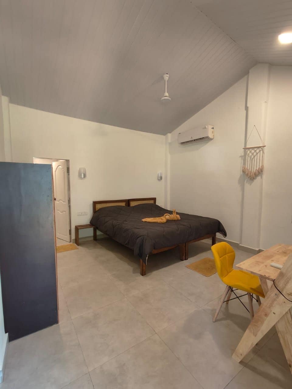 A bedroom with a bed, a small table, a chair, a ceiling fan, and an air conditioning unit.