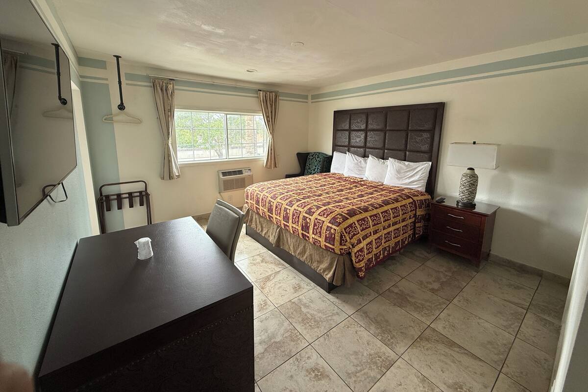 A hotel room with a bed, a desk, a chair, a window with curtains, a wall-mounted TV, and a bedside table with a lamp.