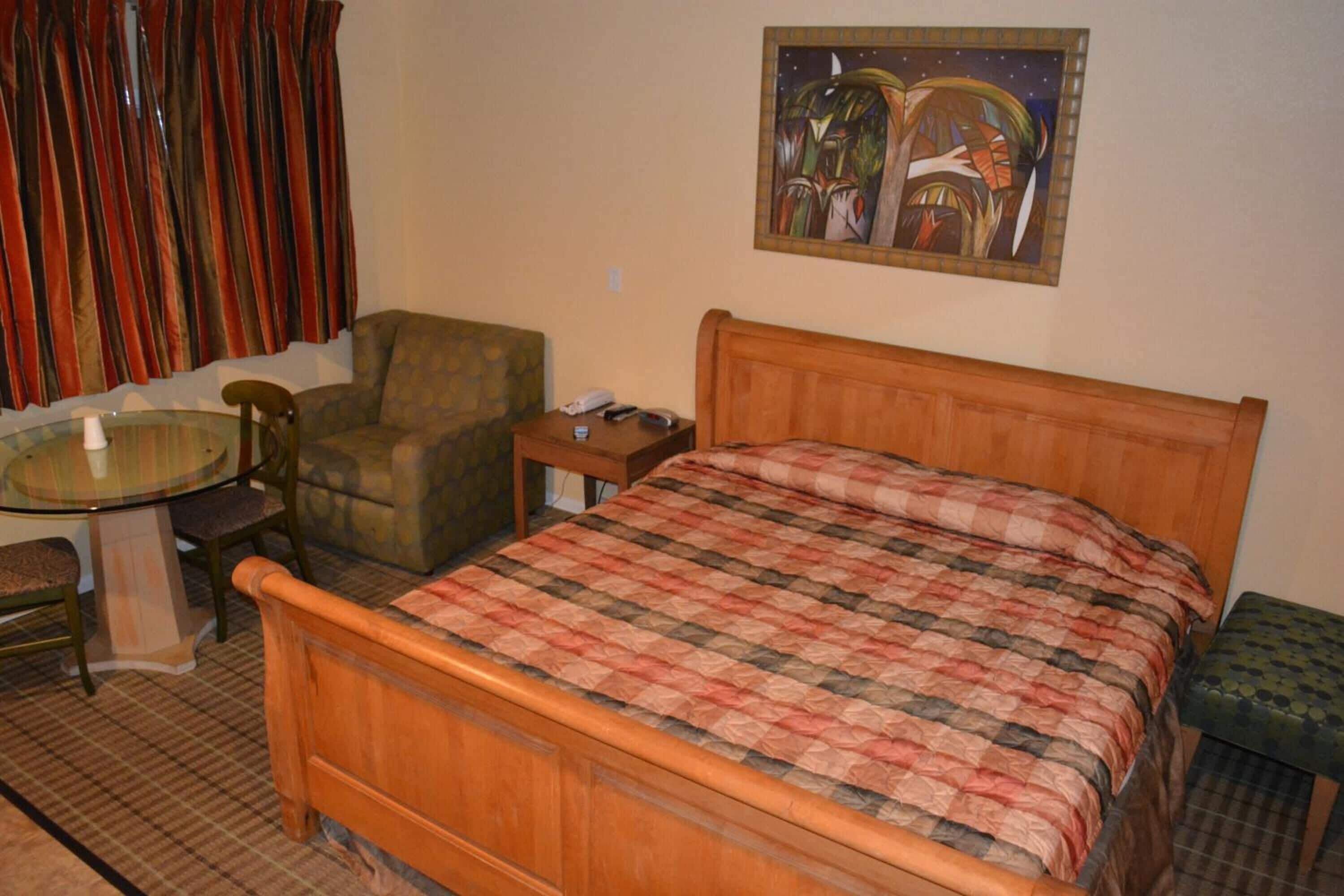 Quarto com cama de madeira, uma mesa redonda com cadeiras, uma pequena escrivaninha e um quadro na parede.