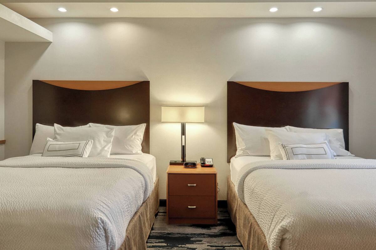 Una habitación de hotel con dos camas, una mesita de noche con lámpara y un cajón pequeño.
