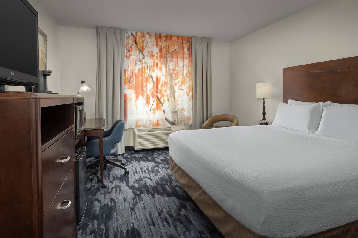 Ein Hotelzimmer mit einem großen Bett, einem Schreibtisch mit Stuhl, einem Fernseher und einem fenster mit Vorhängen.
