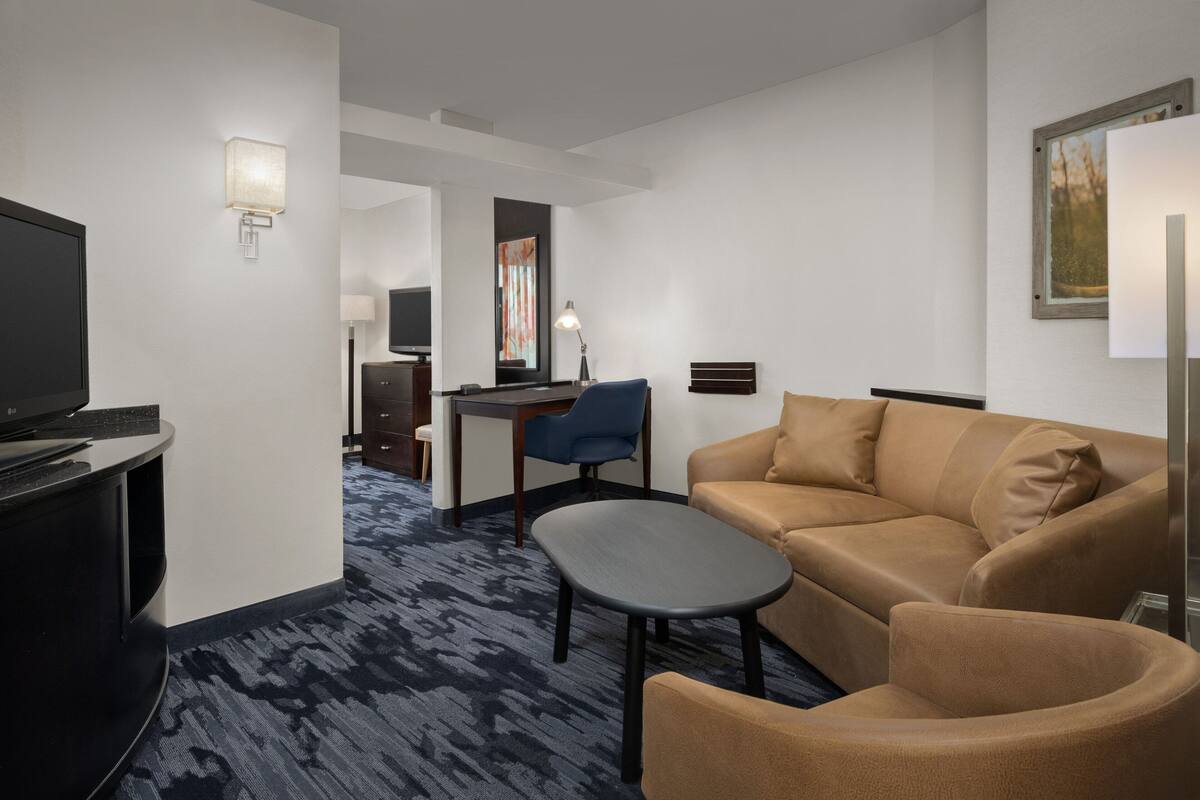 Ein Hotelzimmer mit Sofa, Sesseln und einem kleinen Tisch. Ein Fernseher steht auf einem Schrank, ein Schreibtisch mit Stuhl und eine Lampe sind ebenfalls vorhanden. Der Raum hat einen Teppichboden und ein Bild an der Wand.