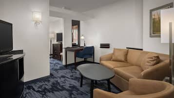 Habitació d'hotel amb sofà, butaca i taula petita. Hi ha una televisió sobre un suport, un escriptori amb una cadira i una làmpada. La habitació té el sòl amb moqueta i un quadre a la paret.