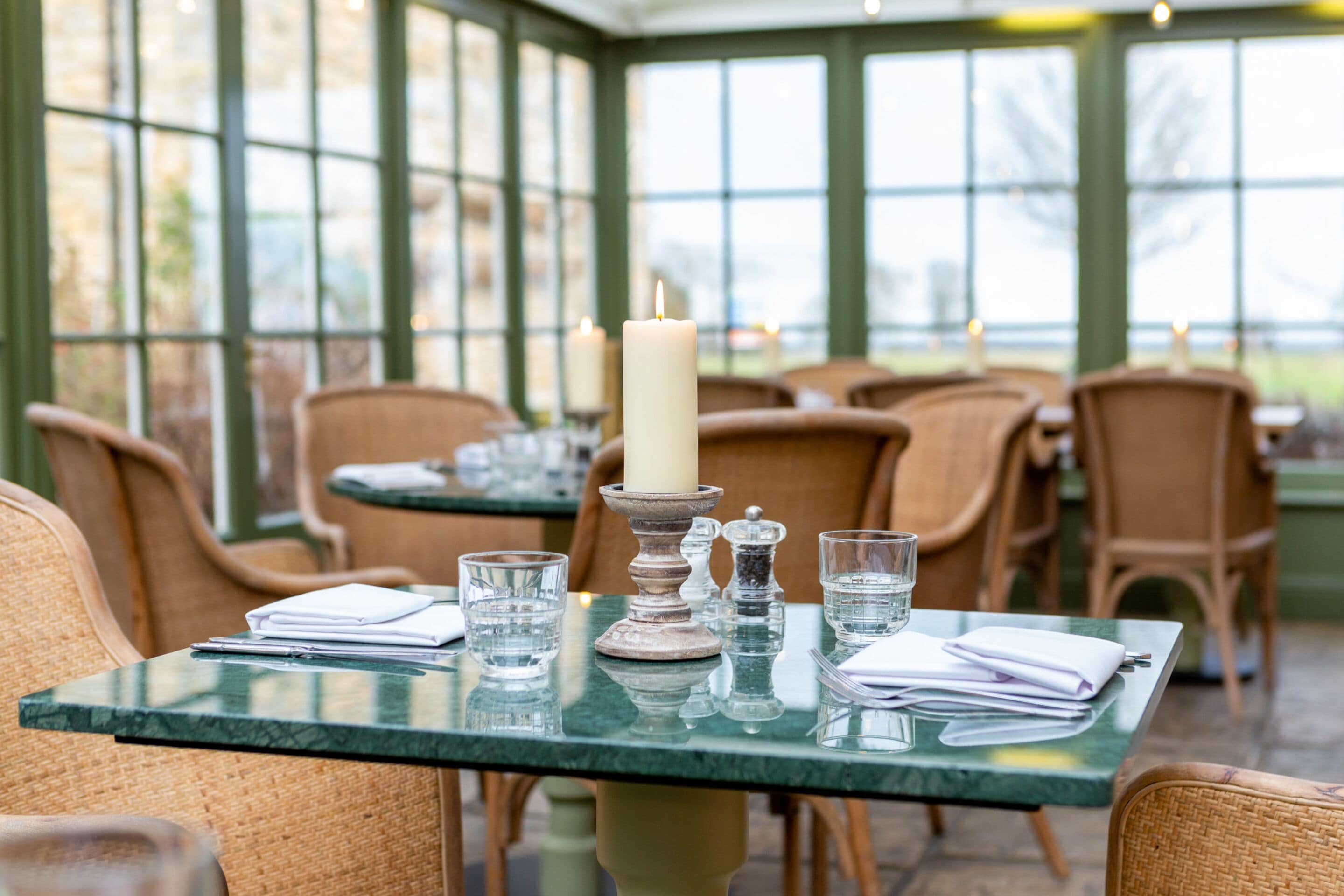 A dining area with tables set for two, featuring green glassware and a candle centerpiece.