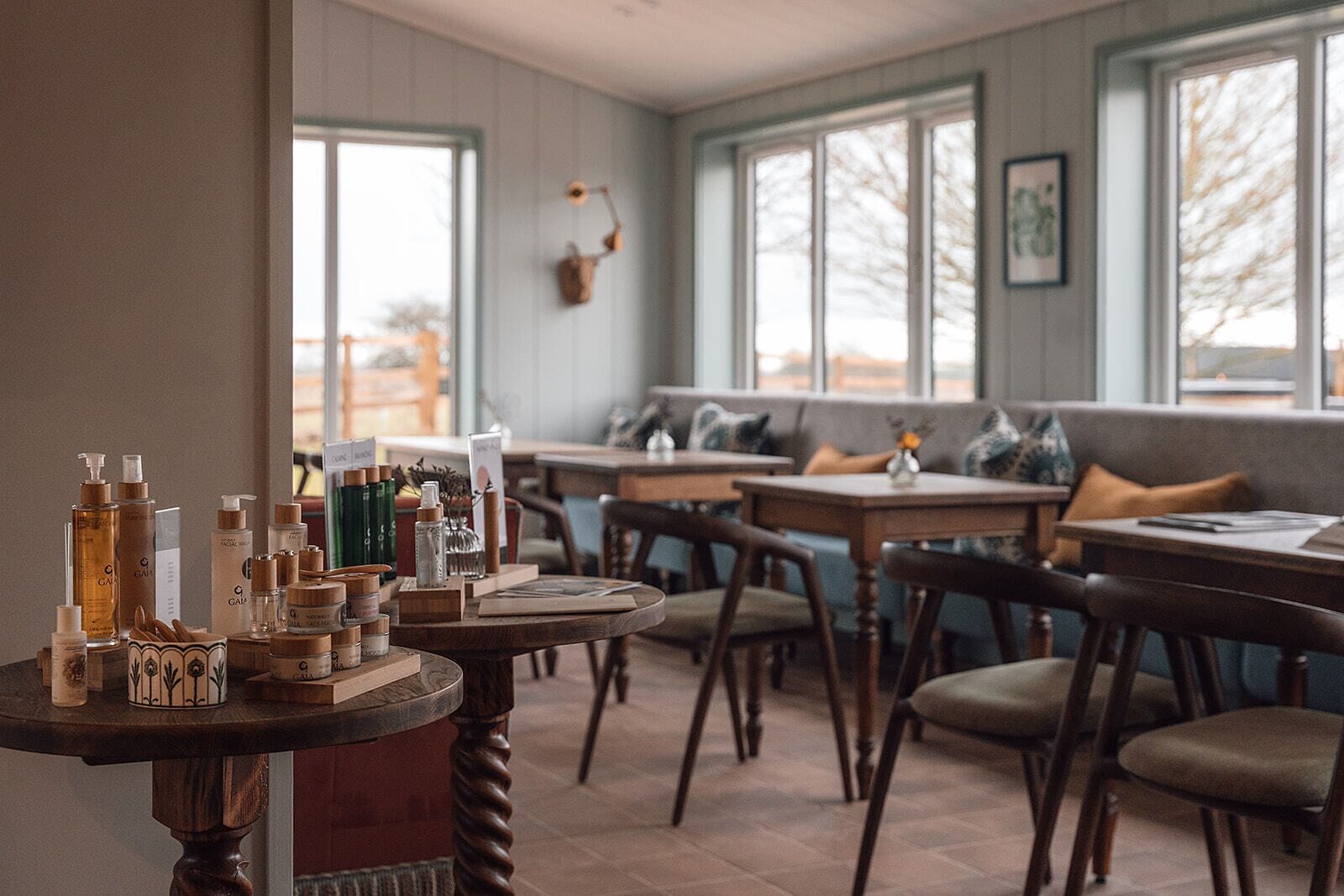 A cozy restaurant interior with wooden tables and chairs, a variety of beverages on a table, and large windows with a view of trees.