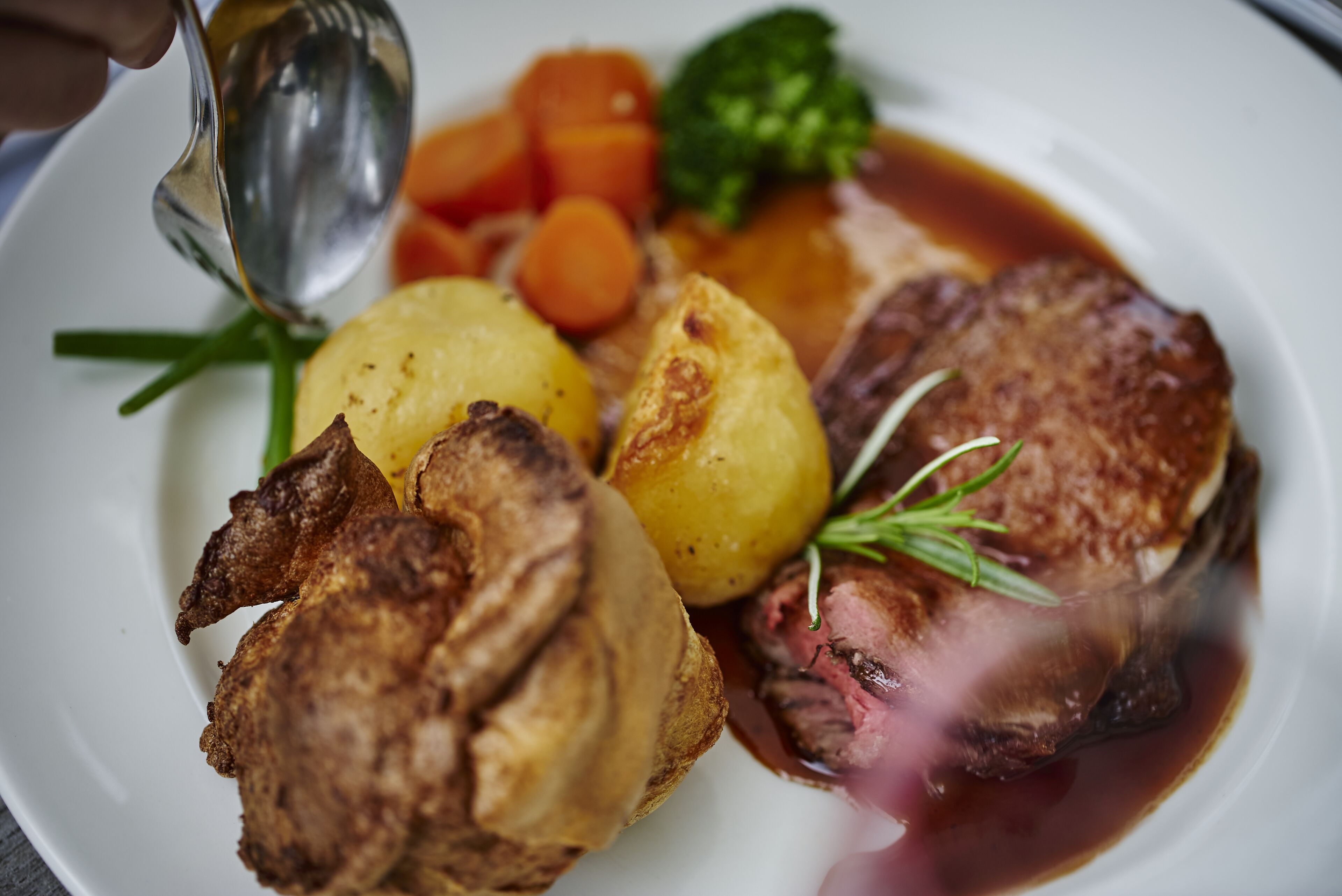 A plate of food with roast beef, potatoes, carrots, and a green vegetable, possibly broccoli, garnished with a sprig of rosemary.