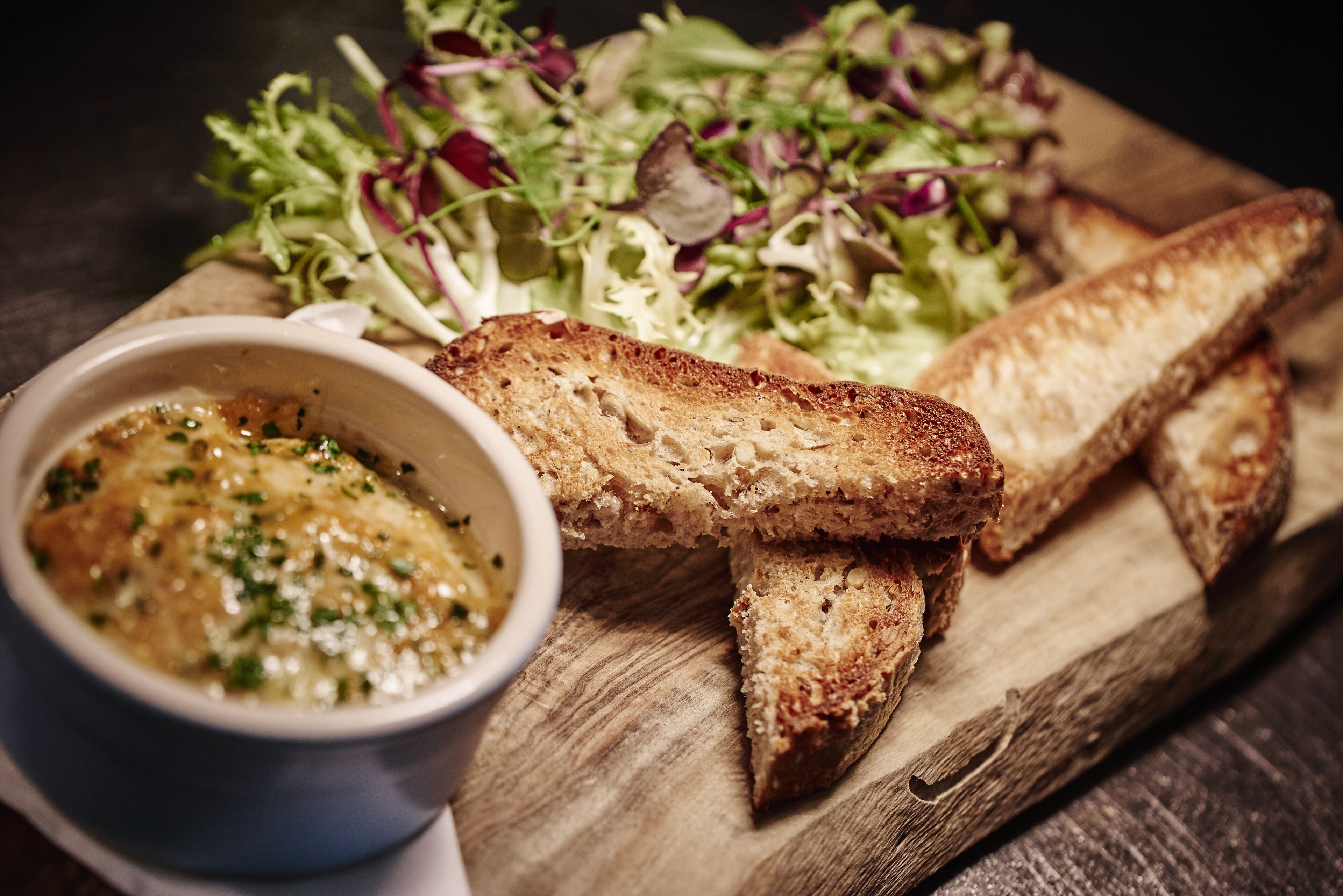 A wooden board with toasted bread, a bowl of sauce, and a mixed green salad.